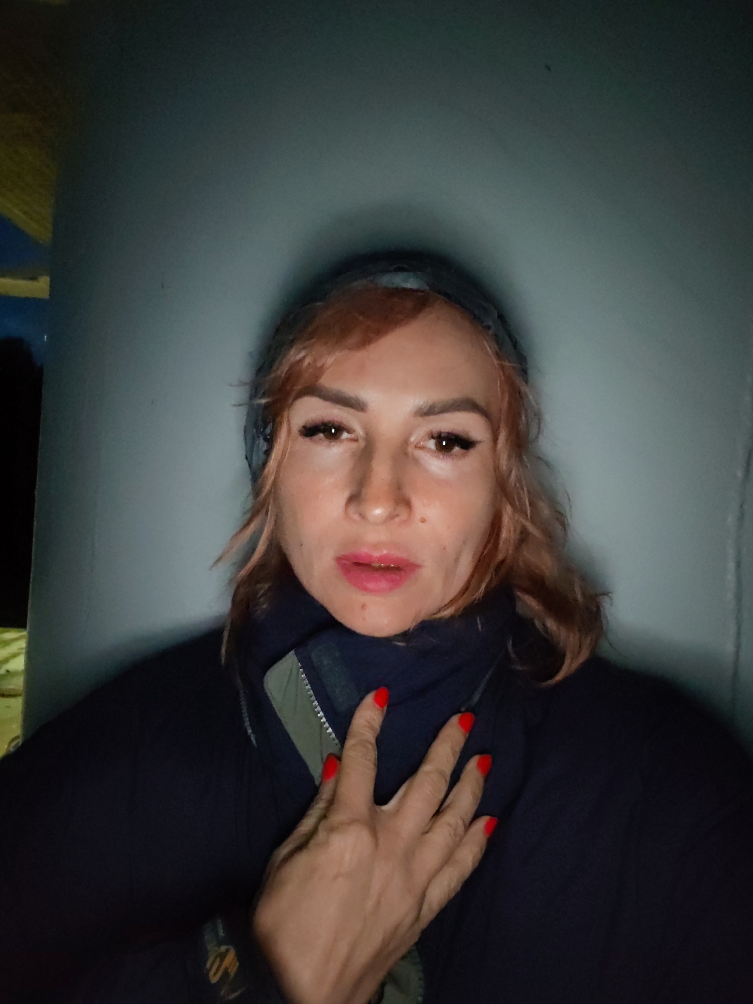 A woman with reddish-brown hair, wearing a dark jacket and a headscarf, is pictured in a close-up shot. She has makeup on, including eyeliner and lipstick. Her left hand, with red nail polish, is placed on her chest. The background appears to be a plain, light-colored wall. The lighting creates a shadow on the wall behind her head. The image was taken outdoors, possibly in the city of Devyatiny, Russia, but this cannot be definitively confirmed from the visual information alone. The overall scene is dimly lit, suggesting it may be evening or nighttime.