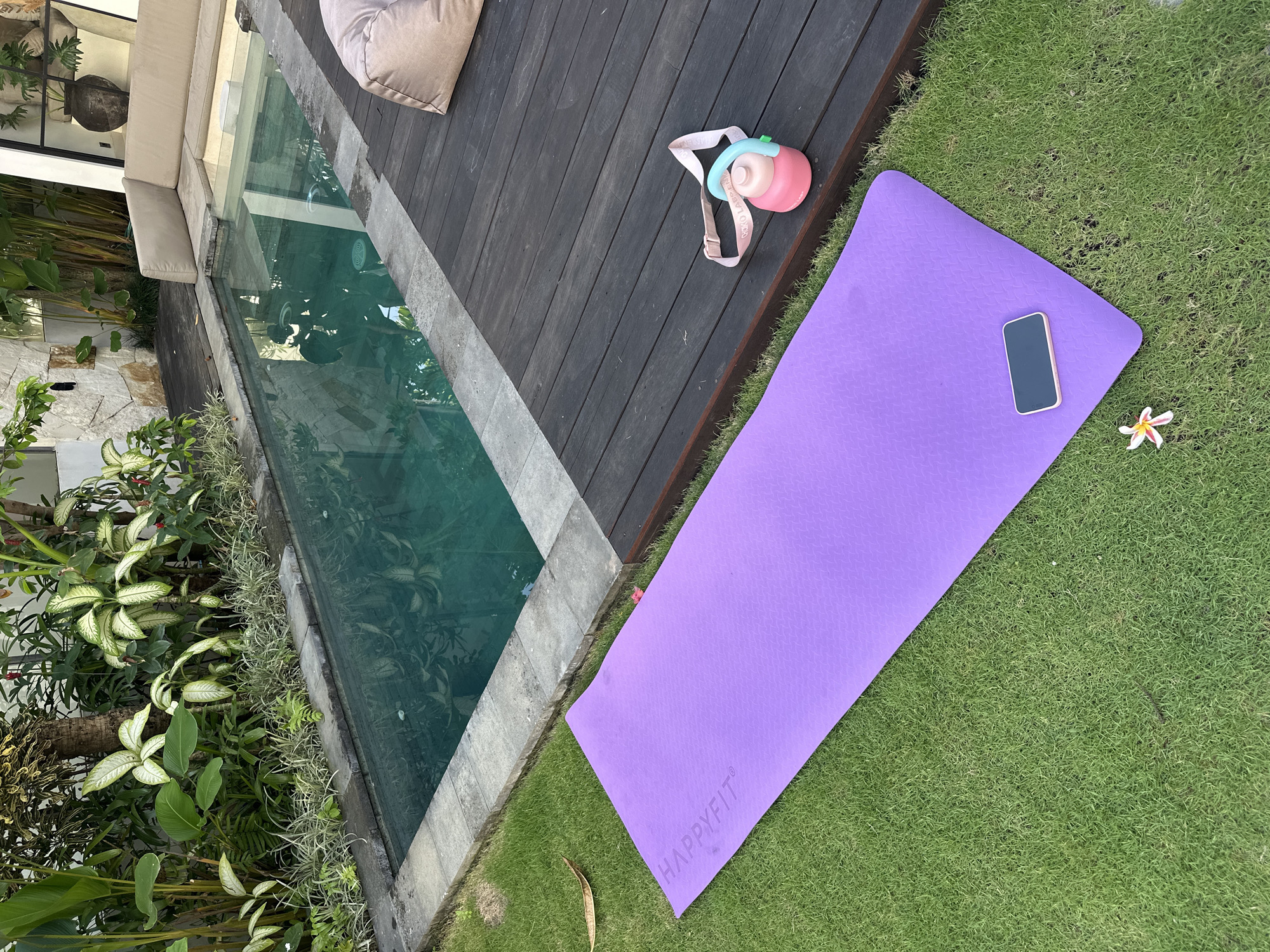 The photograph captures an outdoor scene in North Kuta, Indonesia, featuring a swimming pool area. A purple yoga mat rests on a patch of green grass, with a cell phone placed on top. A water bottle with a pink body and light blue accents lies on a dark wooden deck adjacent to the grass. A white flower is near the yoga mat. The pool is bordered by a stone edge and lush green vegetation. A beige cushion rests on the wooden deck near the pool. A stone wall can be seen with a glass door and potted plant.