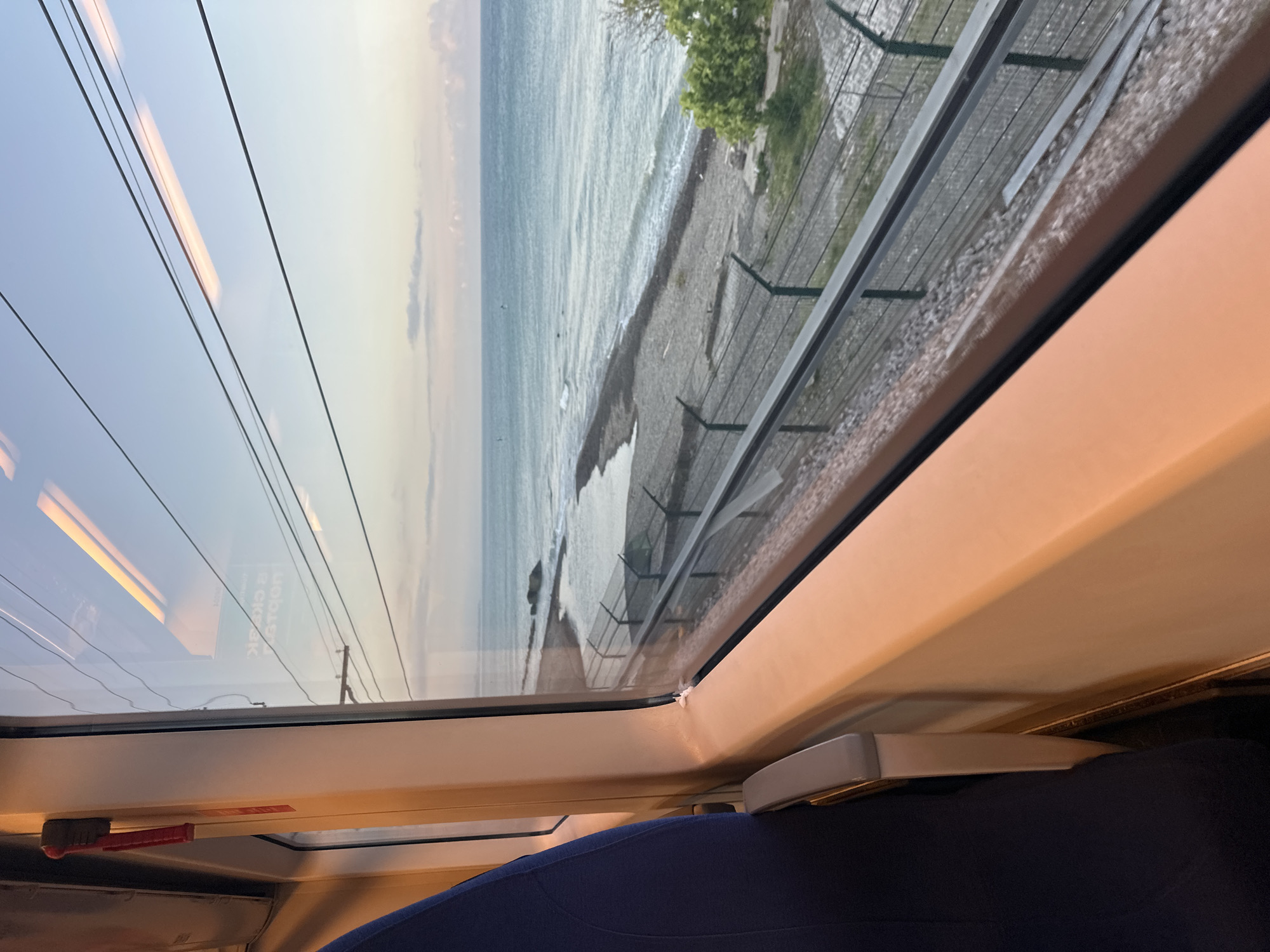 The image is taken from inside a train car, looking out a window at a coastal scene. A dark blue train seat is visible in the foreground on the left. The window frame is a light peachy-orange color. Outside the window, the ocean stretches to the horizon under a pale sky with some clouds. The shoreline is rocky and appears to be a beach. A fence runs along the coastline, parallel to what looks like train tracks. There is some greenery on the right side of the image. Reflections of the train's interior, including light fixtures, are visible on the window.