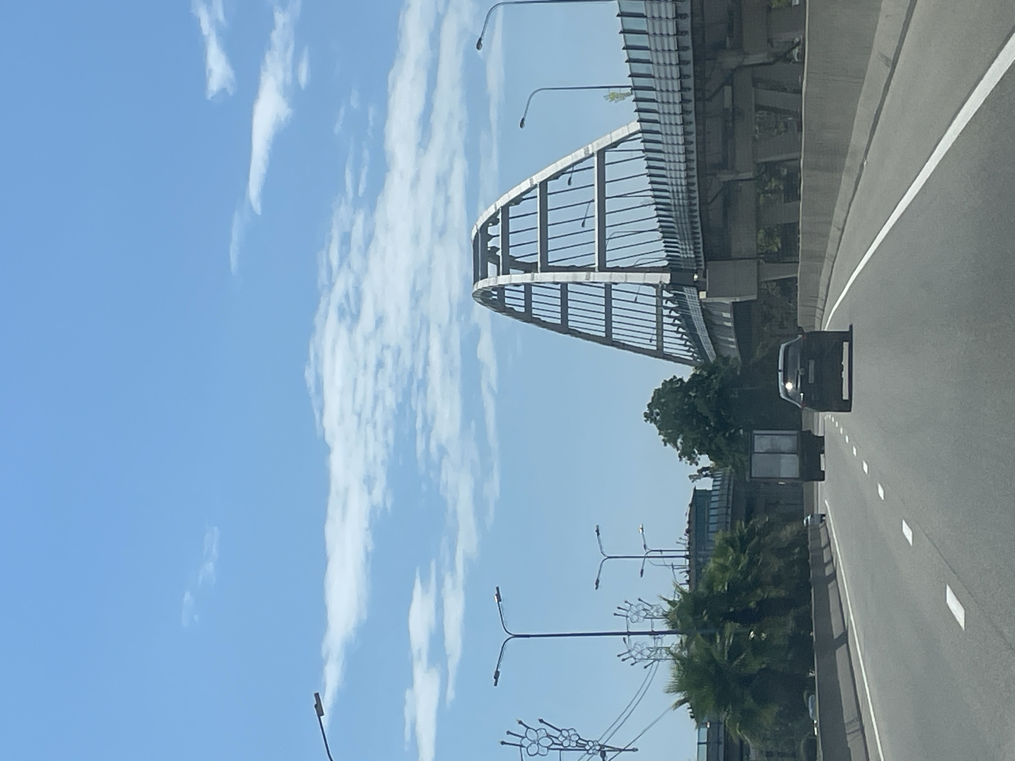 The image shows a view from a car traveling on a highway in Sochi, Russia. In the foreground, the road stretches forward with white lane markings. A black SUV is visible in the right lane, and a white truck is in the left lane. The background features a large, modern arched bridge with a metal lattice structure. Palm trees and other greenery are visible along the side of the highway. The sky is blue with scattered white clouds. The scene appears to be a sunny day.