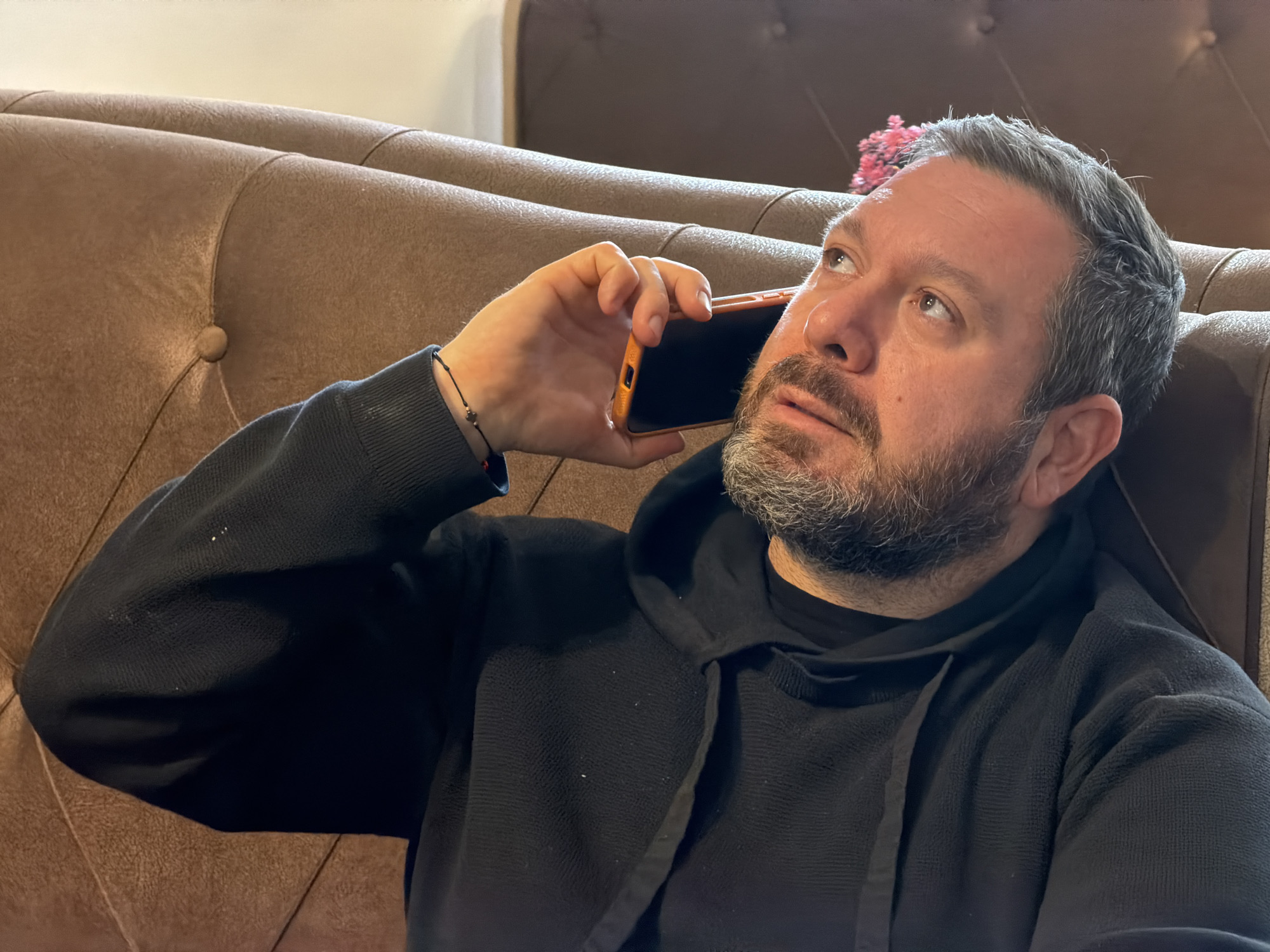 A middle-aged man with graying hair and a beard is sitting on a brown upholstered sofa with button detailing. He is wearing a black hooded sweatshirt and holding an orange-cased smartphone to his ear, looking upwards. He is wearing a bracelet on his left wrist. A possible location is Sofia, Bulgaria.