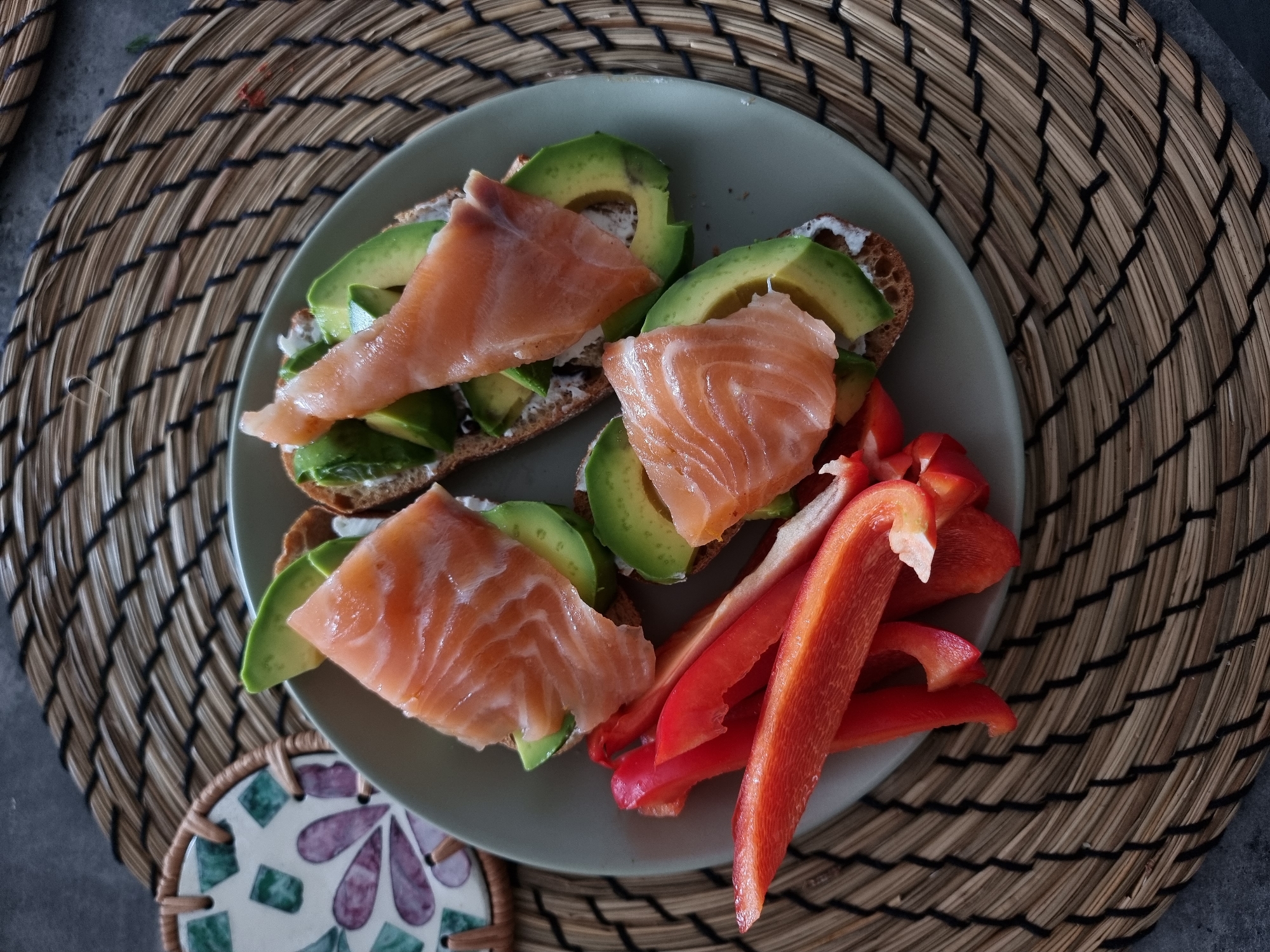 The image depicts a plate of food placed on a woven placemat, likely in a residential setting, as there are no overt markers indicating a specific public location within Ramat Gan, Israel. The plate contains three pieces of toast topped with avocado slices and what appears to be smoked salmon. Adjacent to the toast are several slices of red bell pepper. The food appears to be prepared for consumption. A coaster with a mosaic pattern sits near the plate. The placemat is round and woven from natural fibers. The background is a neutral gray surface.