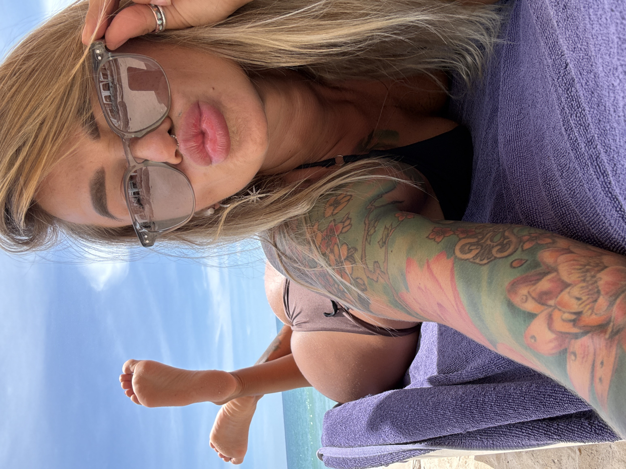 A woman with blonde hair and tattoos on her arm lies on a purple towel on a beach. She is wearing sunglasses, a nose ring, and a brown bikini. Her feet are raised in the air. The ocean and sky are visible in the background. The location is Choeng Thale, Thailand.