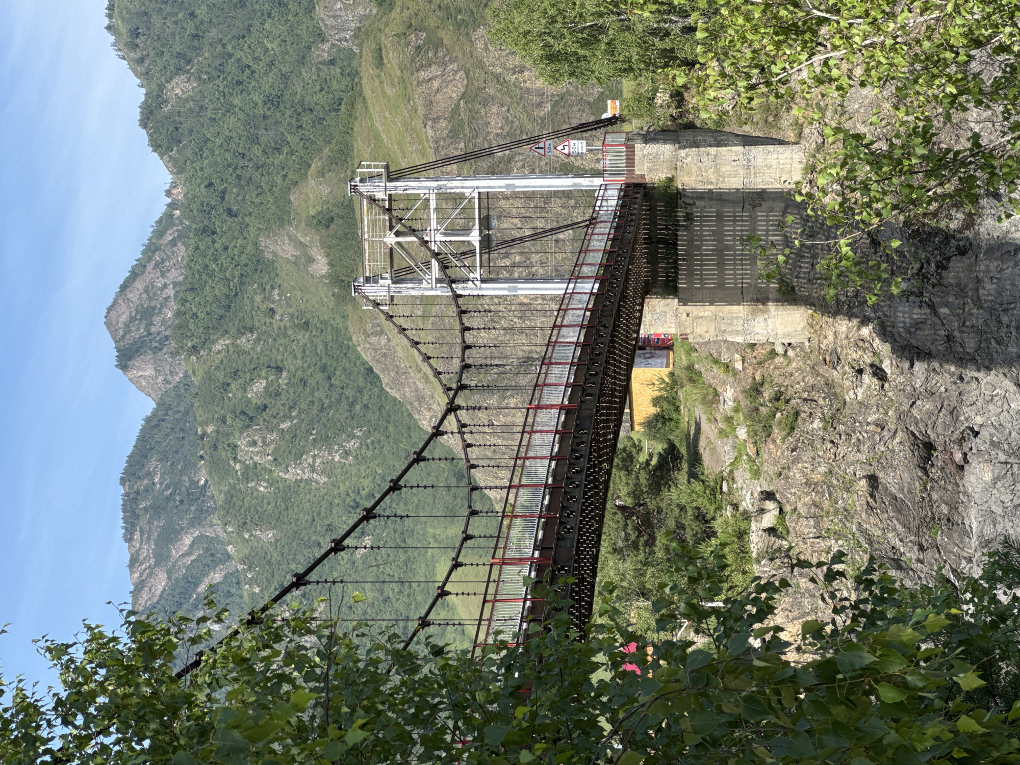 The image shows a suspension bridge crossing a rocky gorge with lush green vegetation. The bridge has a metal deck with red and white railings and is supported by two large concrete piers and suspended by thick cables attached to tall white towers. The surrounding landscape is mountainous, with steep, tree-covered slopes under a clear blue sky. On the right side of the bridge, there are two road signs, one indicating a 50-meter distance and another with arrows and distances of 50m and 400m. The lower part of the scene reveals a rocky terrain and foliage. The overall scene depicts a natural landscape with a prominent man-made structure, suggesting a location like Edigan, Russia, known for its scenic mountainous terrain and bridges.