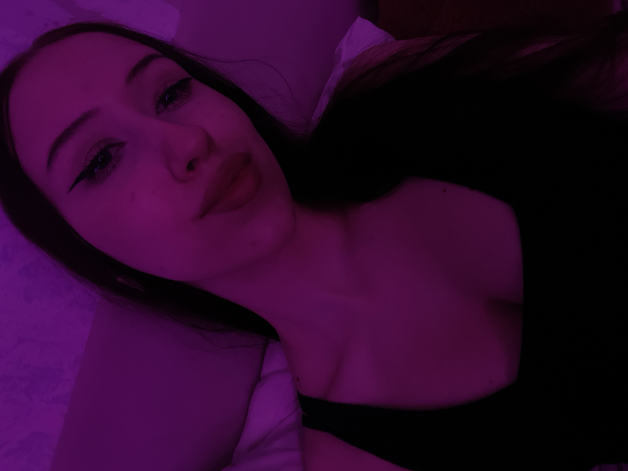The image shows a young woman with long dark hair, wearing a black top. She has on eyeliner and lip gloss. The lighting in the image is purple. The woman appears to be indoors, lying down on a surface that is either a bed or a couch. The background is dimly lit and appears to be a wall with a textured surface. There is no indication of the woman's location within the city of Votkinsk, Russia, as the image does not contain any recognizable landmarks or identifying features of that city.