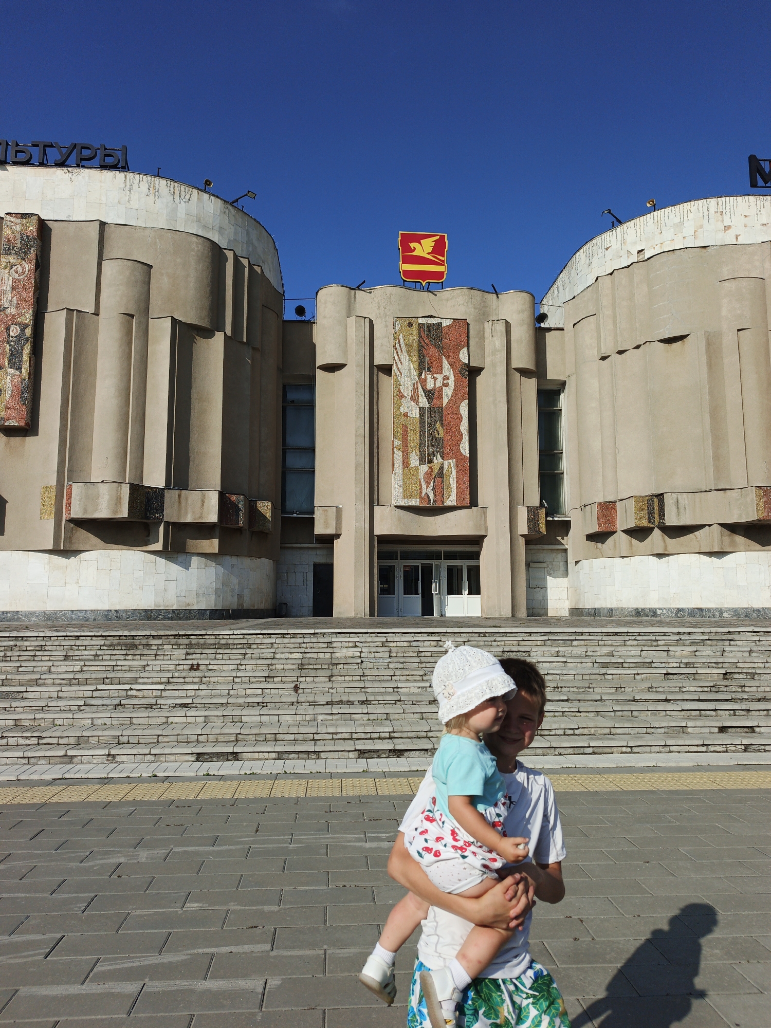 The image depicts a bright, sunny day with a clear blue sky over the Palace of Culture in Zlatoust, Russia. The imposing building features a textured beige facade with decorative mosaic panels and a prominent red and yellow emblem on its roof. A grand staircase leads up to the building's entrance. In the foreground, a young boy is carrying a toddler, who is wearing a white hat and a blue and white cherry-patterned dress. Both children are smiling. The ground in front of the building is paved with grey interlocking stones and a yellow tactile paving strip.