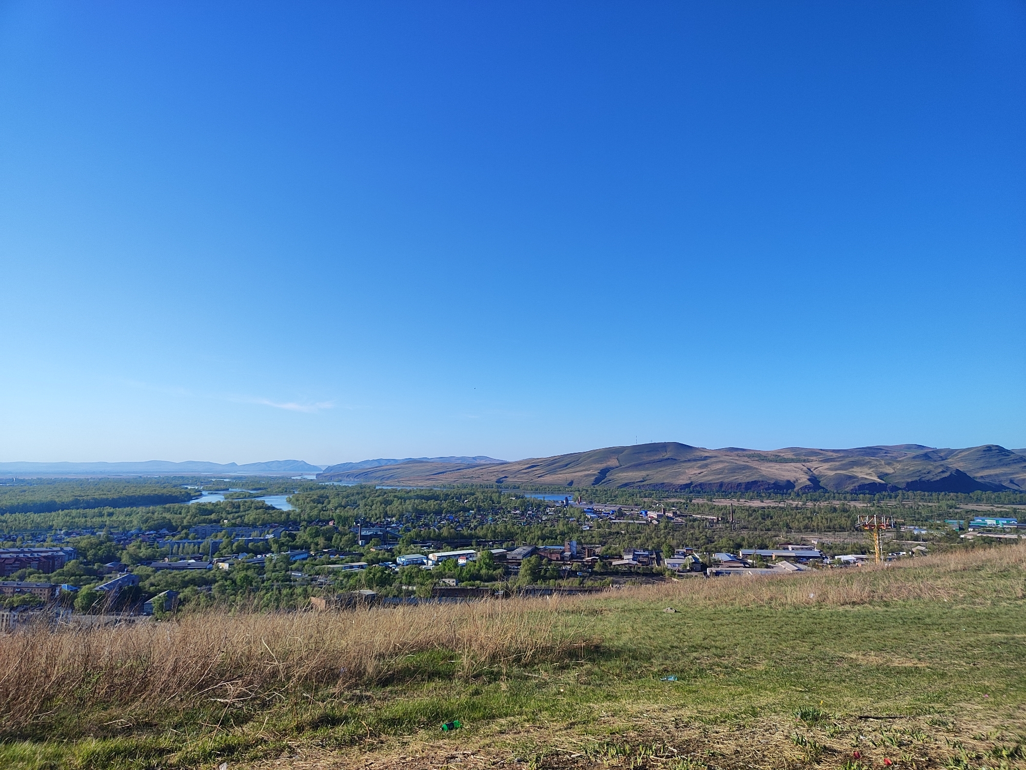 A wide-angle shot captures a landscape of Unknown, Unknown, from an elevated perspective. The foreground is dominated by a grassy hill, transitioning from dry, brown grass to patches of green. In the middle ground, a town or city is visible, characterized by a mix of buildings interspersed with greenery. Beyond the town, a body of water meanders through the scene, surrounded by lush forests. The background features rolling hills, their slopes covered with sparse vegetation. The sky is a clear, cloudless blue.
