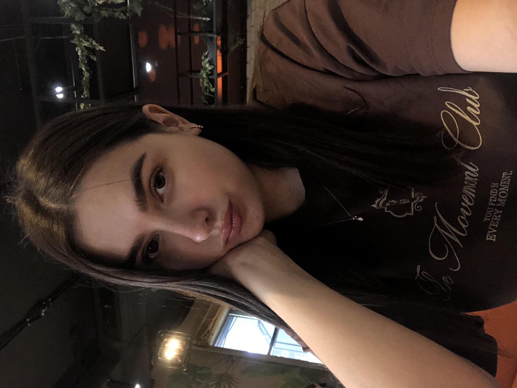 The image shows a close-up selfie of a young woman with long, dark brown hair and a brown t-shirt. She is resting her chin on her hand and looking towards the camera with a slightly pensive expression. She is wearing a delicate necklace with a small pendant and small hoop earrings. The background is dimly lit and appears to be an indoor setting, possibly a cafe or restaurant, with some greenery and lighting fixtures visible. There is a window on the left side of the frame. Based on the provided information, the image was taken in Izhevsk, Russia.