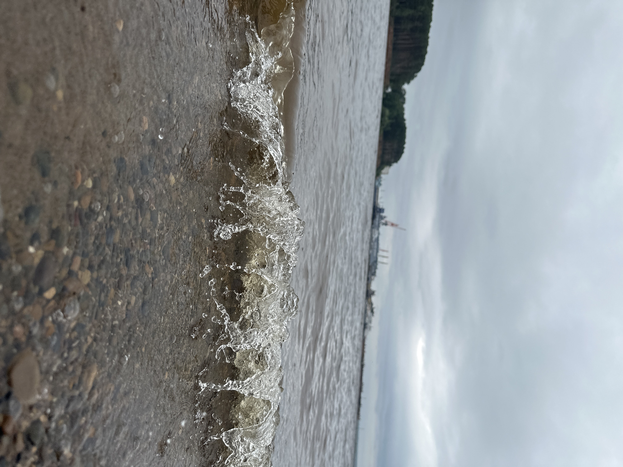 A close-up, vertical shot captures a wave breaking on a pebbly shore. The foreground is dominated by the wet sand and stones, with the translucent water of the wave cresting and splashing. Beyond the immediate shore, the water extends to the horizon. In the distance, a tree-covered hillside rises, and further still, structures that appear to be industrial or port facilities are visible, silhouetted against a cloudy sky. The overall mood is calm and natural, with the focus on the dynamic interaction of water and land. The lighting suggests it is daytime, possibly overcast, given the muted tones and soft light. No text is visible in the image.
