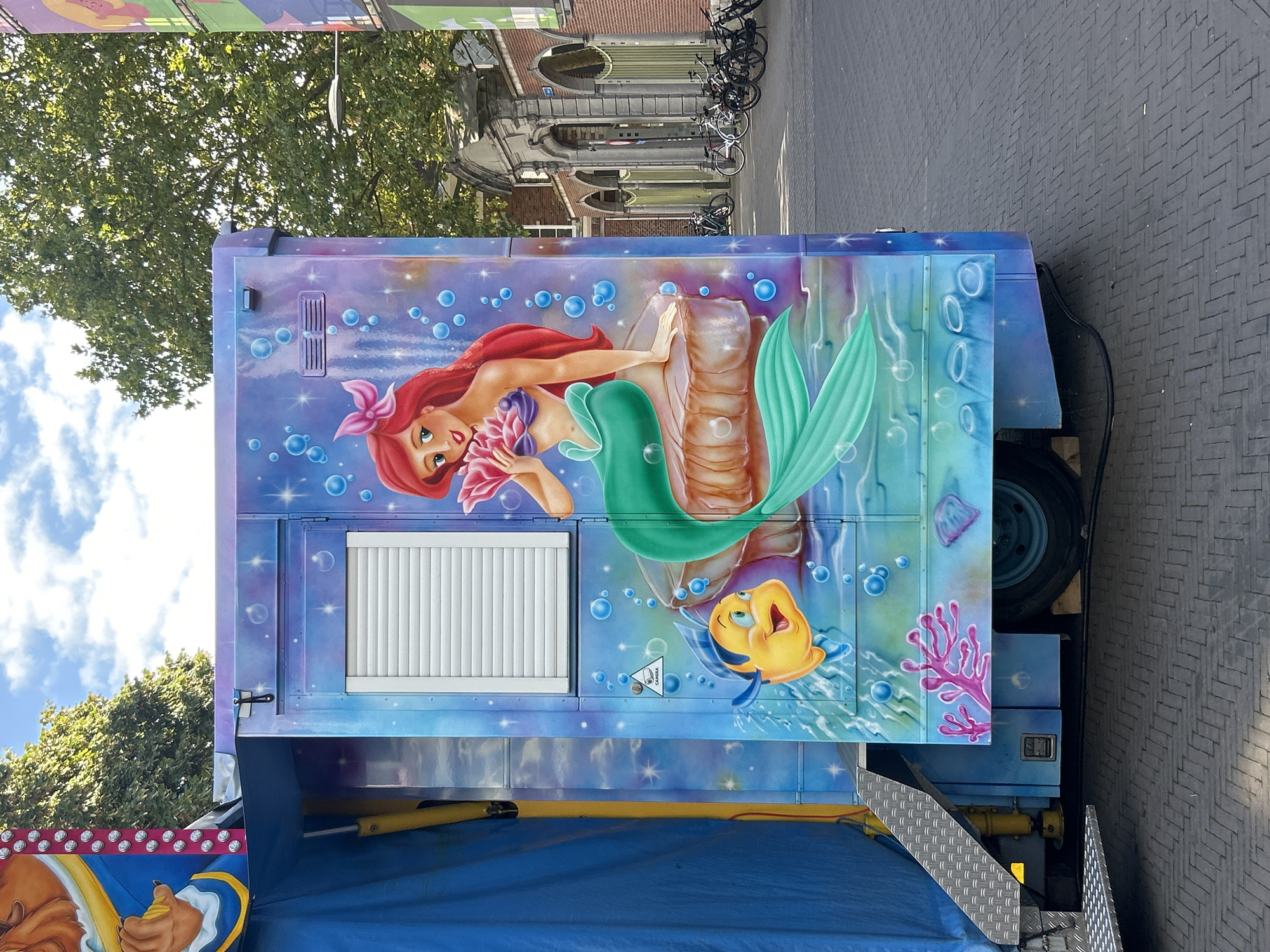 The image shows a brightly painted trailer with a mural of Ariel from Disney's "The Little Mermaid" and her friend Flounder. Ariel is depicted sitting on a rock, holding a pink seashell, with her iconic red hair and green mermaid tail. Flounder, a yellow and blue fish, swims nearby. The trailer has a blue tarp on the left side and a closed window with white blinds. A security camera sticker is visible on the side of the trailer. In the background, there are trees, a building with arches, and several parked bicycles. The scene appears to be on a paved street. This image was taken on the Tiensestraat in Leuven, Belgium, in front of the KADOC building.