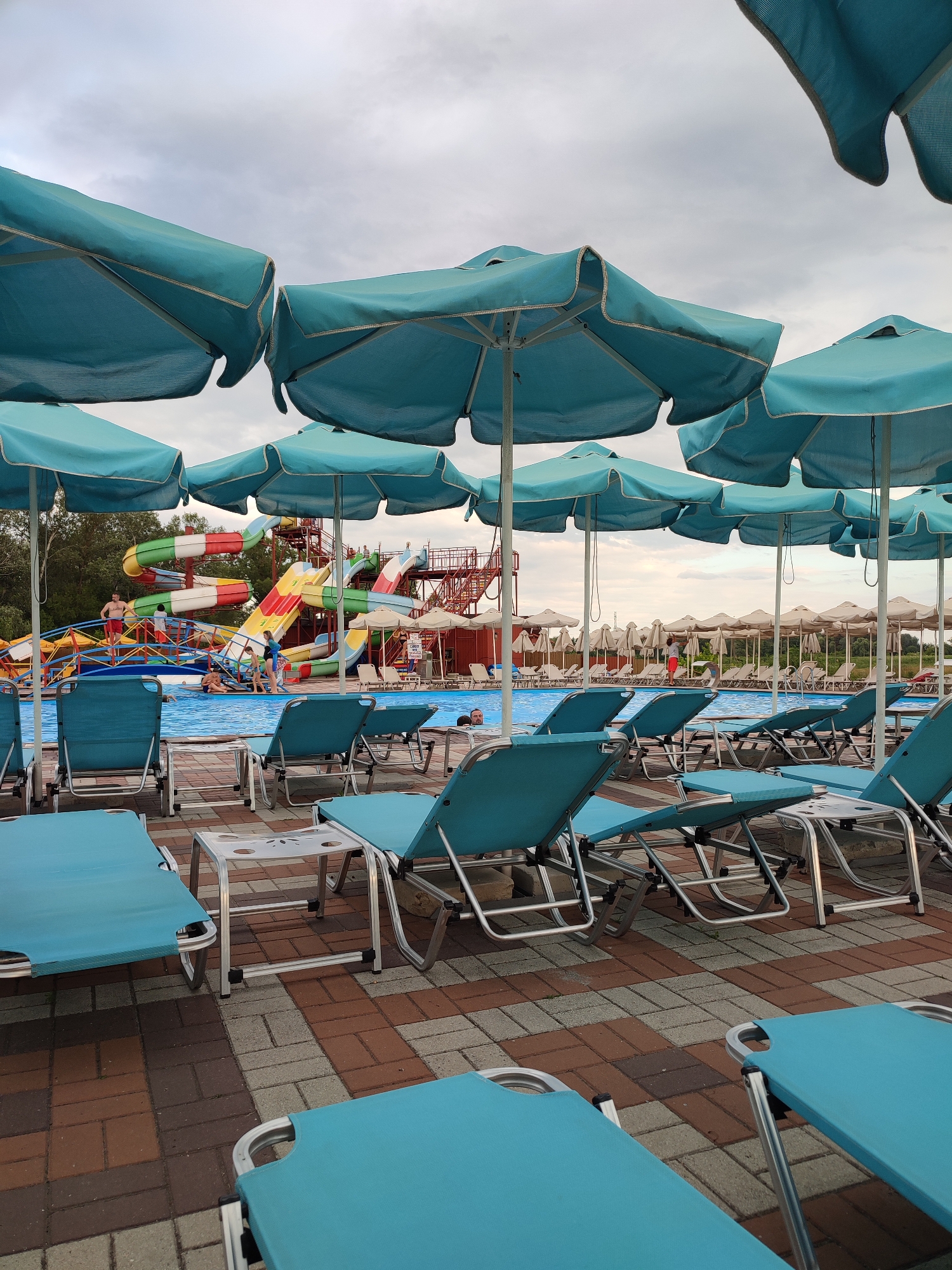 The image depicts an outdoor water park in Shcherbani, Ukraine, under an overcast sky. In the foreground, numerous turquoise lounge chairs with metal frames are arranged on a brick-patterned patio. Several of these chairs are empty, while a few appear to be occupied by unseen individuals. Large, turquoise umbrellas are positioned over some of the chairs, providing shade. Beyond the patio, a bright blue swimming pool is visible, with several people swimming and relaxing in the water.
In the background, a colorful water slide complex dominates the scene. The slides feature various colors, including red, yellow, green, and blue, and are supported by a multi-level metal structure. Some individuals are seen on the platforms and stairs of the water slide complex, seemingly waiting to use the slides or observing others. To the right of the water slides, rows of smaller, light-colored umbrellas are set up, with more empty lounge chairs beneath them. Trees and greenery are visible in the distance, behind the water park structures. The overall atmosphere suggests a recreational area that is either in its early stages of activity or experiencing a lull in patronage due to the weather or time of day.
