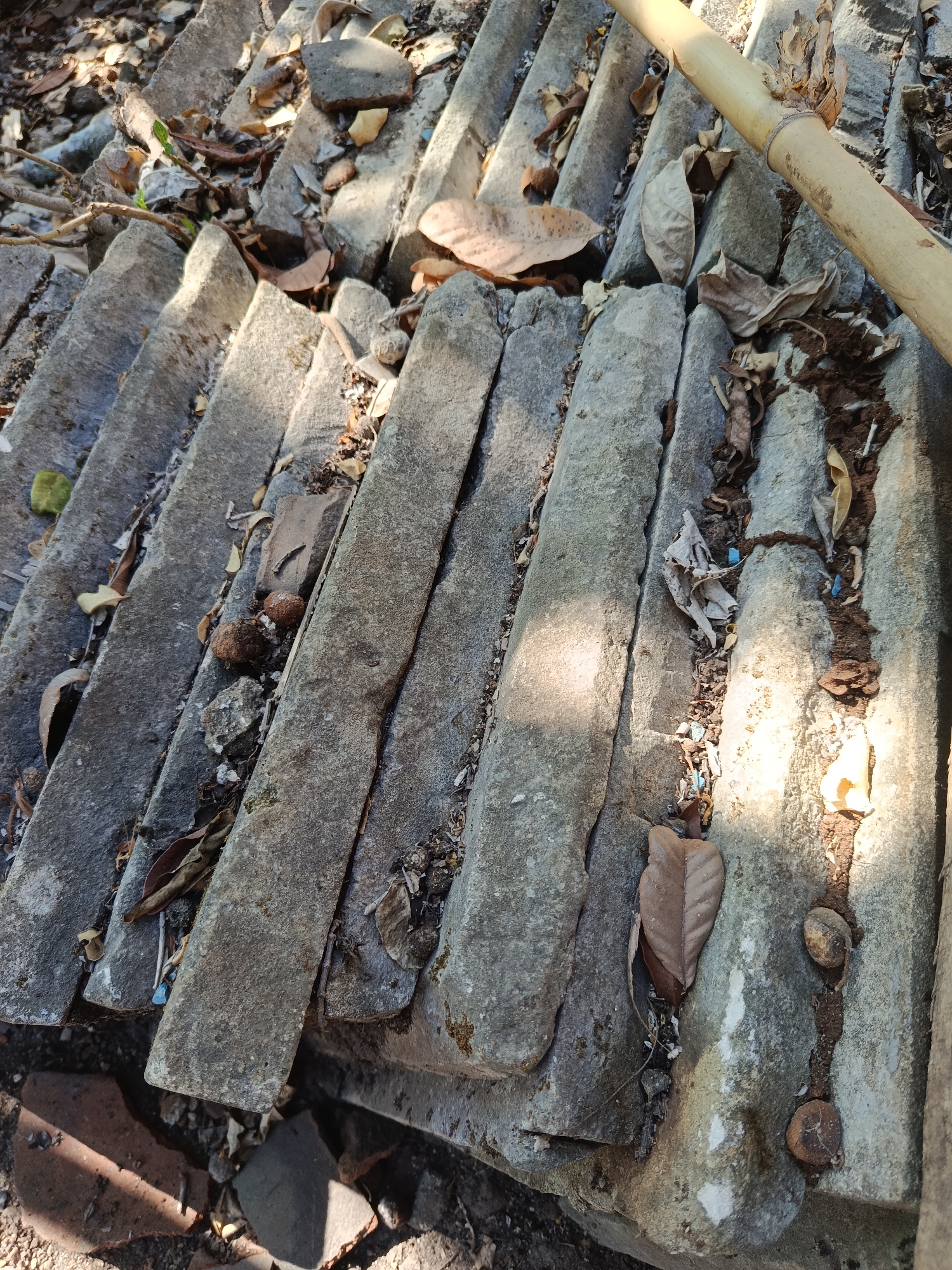 The image shows a close-up, overhead view of weathered, gray concrete roof tiles, likely part of an older structure, possibly in Karangmojo, Indonesia, based on the prompt's context. The tiles are arranged in parallel rows, with significant gaps between them, filled with dry leaves, dirt, small stones, and debris. A light-colored bamboo pole runs diagonally across the upper right portion of the frame, resting on the tiles. Shadows from unseen objects cast diagonal stripes of light and dark across the textured surface of the roof. The overall impression is one of neglect and natural weathering. There are no people visible in the image.