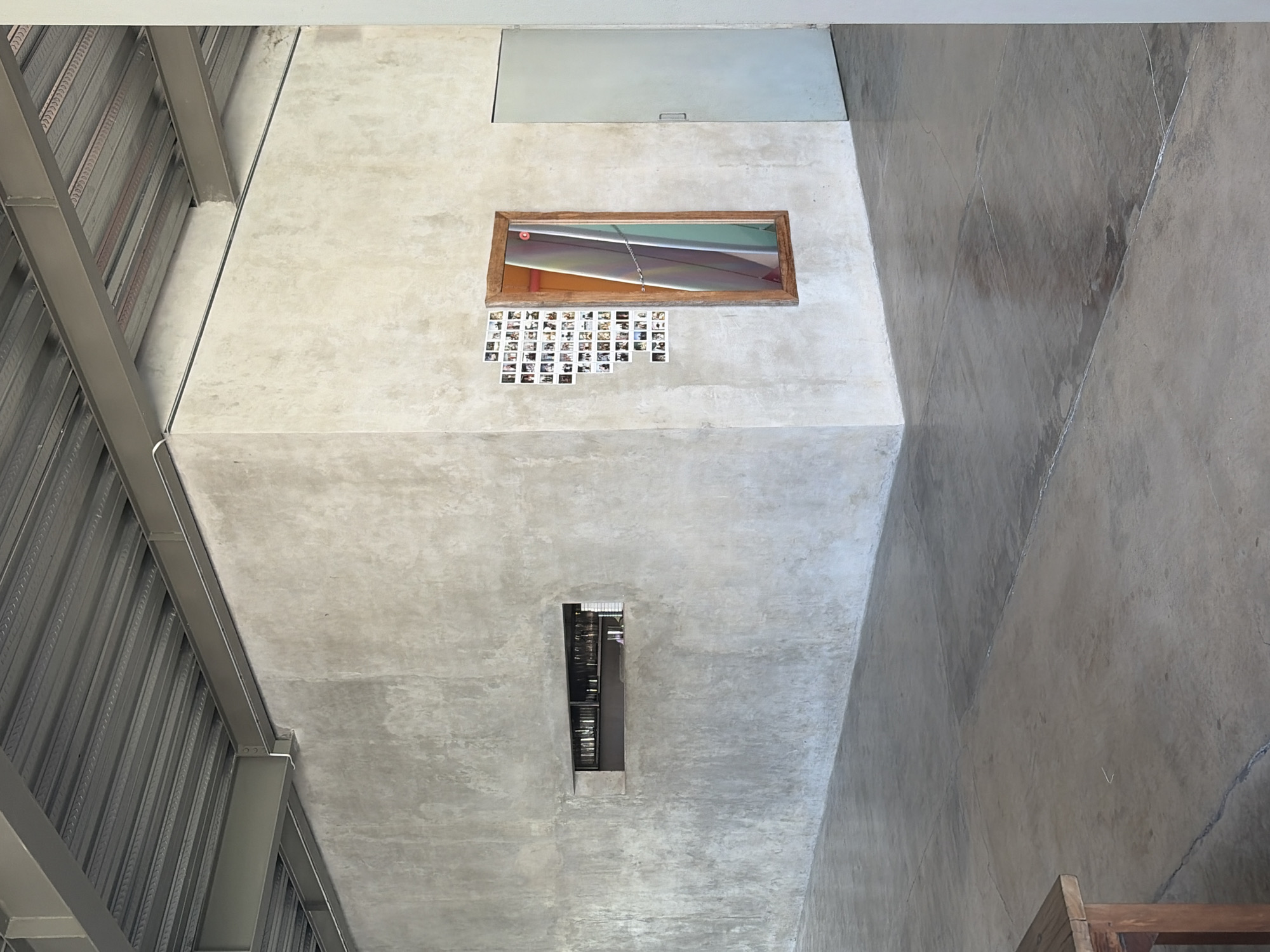 The image shows the interior of a modern building with exposed concrete walls and a corrugated metal ceiling. In the foreground, a polished concrete floor reflects the ambient light. To the left, a wooden table leg is visible. The main wall features a horizontal, recessed shelf filled with what appear to be bottles. To the right of the shelf, a collection of small photographs is arranged in a triangular pattern on the wall, next to a tall, rectangular mirror with a wooden frame. The mirror reflects a surfboard with colorful stripes. Adjacent to the mirror is a closed, pale gray door. The scene is devoid of people. The overall aesthetic is minimalist and industrial. This is the interior of a building in the city of Mengwi, Indonesia.