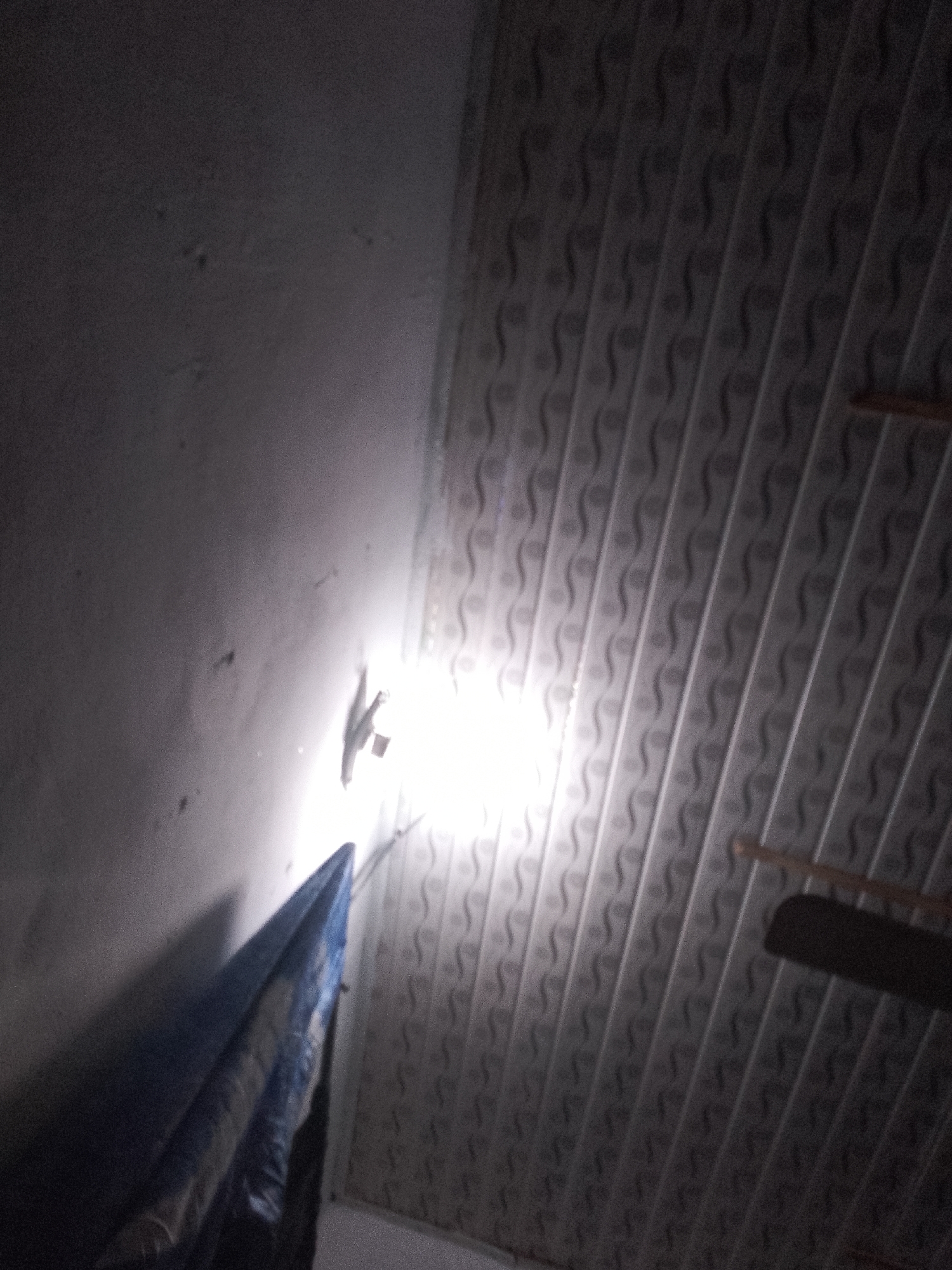 The image shows a dimly lit interior scene. On the left, a textured, light-colored wall is visible, with some dark spots. A blue and white object, possibly a tarp or fabric, is draped downwards from the upper left towards the bottom left. To the right of the wall, a bright light source illuminates the scene, casting a shadow. A small, dark object, possibly an insect or gecko, is seen clinging to the wall near the light. The ceiling, which covers the upper portion of the image, is white and features a repeating patterned design of curved lines and circles. A wooden fan blade is partially visible in the upper right corner, extending from the ceiling. The lighting suggests that the photograph was taken at night or in a poorly lit environment.