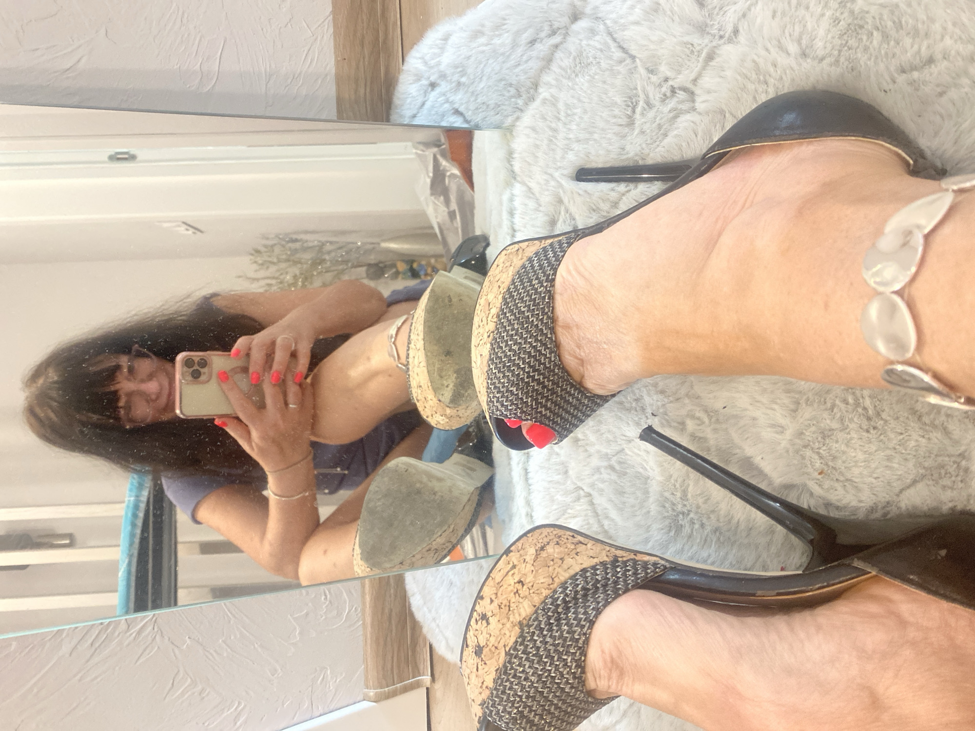 This media file is a selfie taken in a mirror, showing a person's legs and feet adorned with high-heeled shoes. The subject, a person with long, dark hair, is wearing a blue shirt and what appears to be a swimsuit. They are holding a smartphone, presumably to take the photo. The shoes are platform wedges with a cork sole and a black heel strap. The background shows a fluffy rug and a wall with a textured finish. The lighting suggests an indoor setting, possibly a bedroom or dressing area. The image appears to be taken during the day. There is no visible text in the image. The location context provided is Novi Petrivtsi, Ukraine, but this information is not visually discernible from the image itself.
