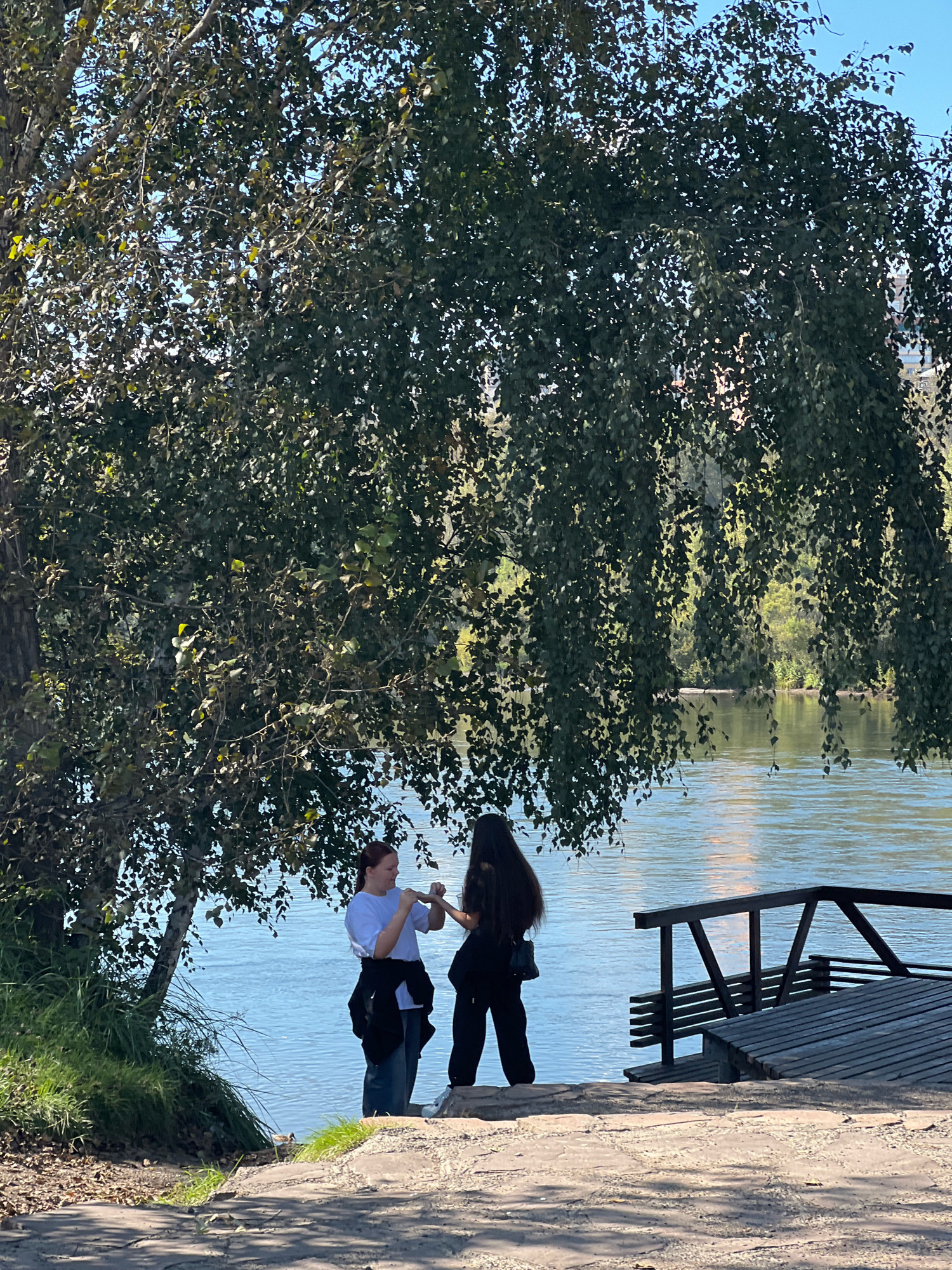 Two women are standing by a body of water. One woman is wearing a white t-shirt and blue jeans, and the other is wearing a black t-shirt and black pants. They are standing on a stone path near a wooden deck. A large tree with green leaves is overhead, casting shadows. The body of water appears to be a river. In the background, trees and buildings are visible. This scene is consistent with the waterfront area of the Yenisei River in Krasnoyarsk, Russia.