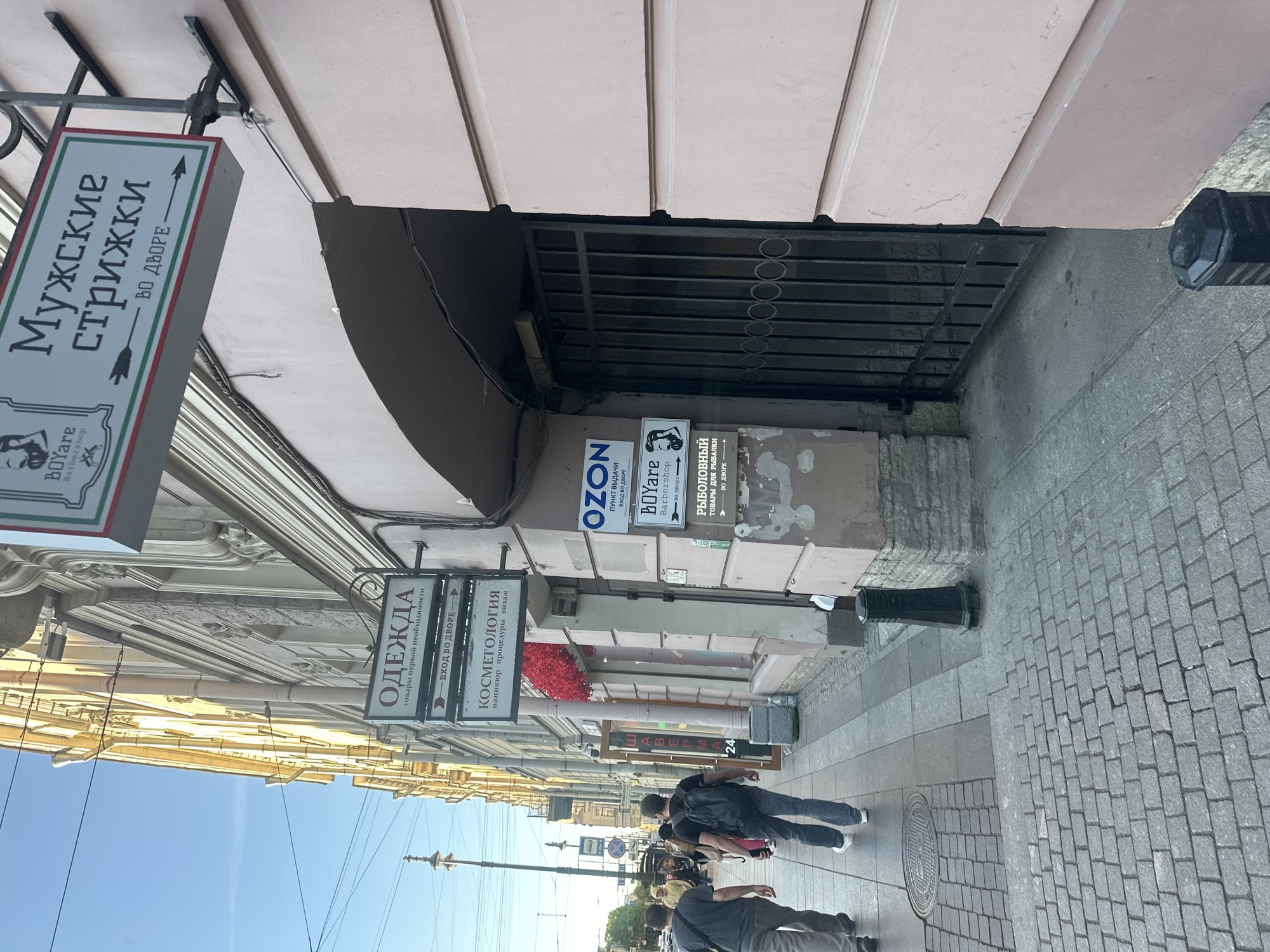 The image shows a street scene in a city. A pale pink building with cream-colored trim lines the right side of the frame, featuring multiple signs advertising various businesses. One sign, in green and white, reads "МУЖСКИЕ СТРИЖКИ" (Men's Haircuts) with an arrow indicating "ВО ДВОРЕ" (In the courtyard). Below this, a smaller sign depicts a barber's pole and text "BOYare Barbershop". Further down the building, another sign in blue and white says "OZON" with "пункт выдачи" (pickup point) and "вход во дворе" (entrance in the courtyard). There is a large black metal gate leading into a courtyard.
On the left side of the image, a sidewalk is visible with people walking. Two men are walking away from the camera. The man on the left wears a dark t-shirt and grey pants. The man on the right wears a black t-shirt and dark jeans with a backpack. The sidewalk is paved with grey stone blocks and lighter grey rectangular tiles. Further down the sidewalk, a sign with Cyrillic text reads "ОДЕЖДА" (Clothing) and "товары первой необходимости" (essential goods), with an arrow also pointing "вход во дворе". Another sign below it reads "КОСМЕТОЛОГИЯ" (Cosmetology) with services listed.
The buildings in the background are yellow and appear to be several stories high. The sky is clear and blue. The overall environment suggests a commercial street.