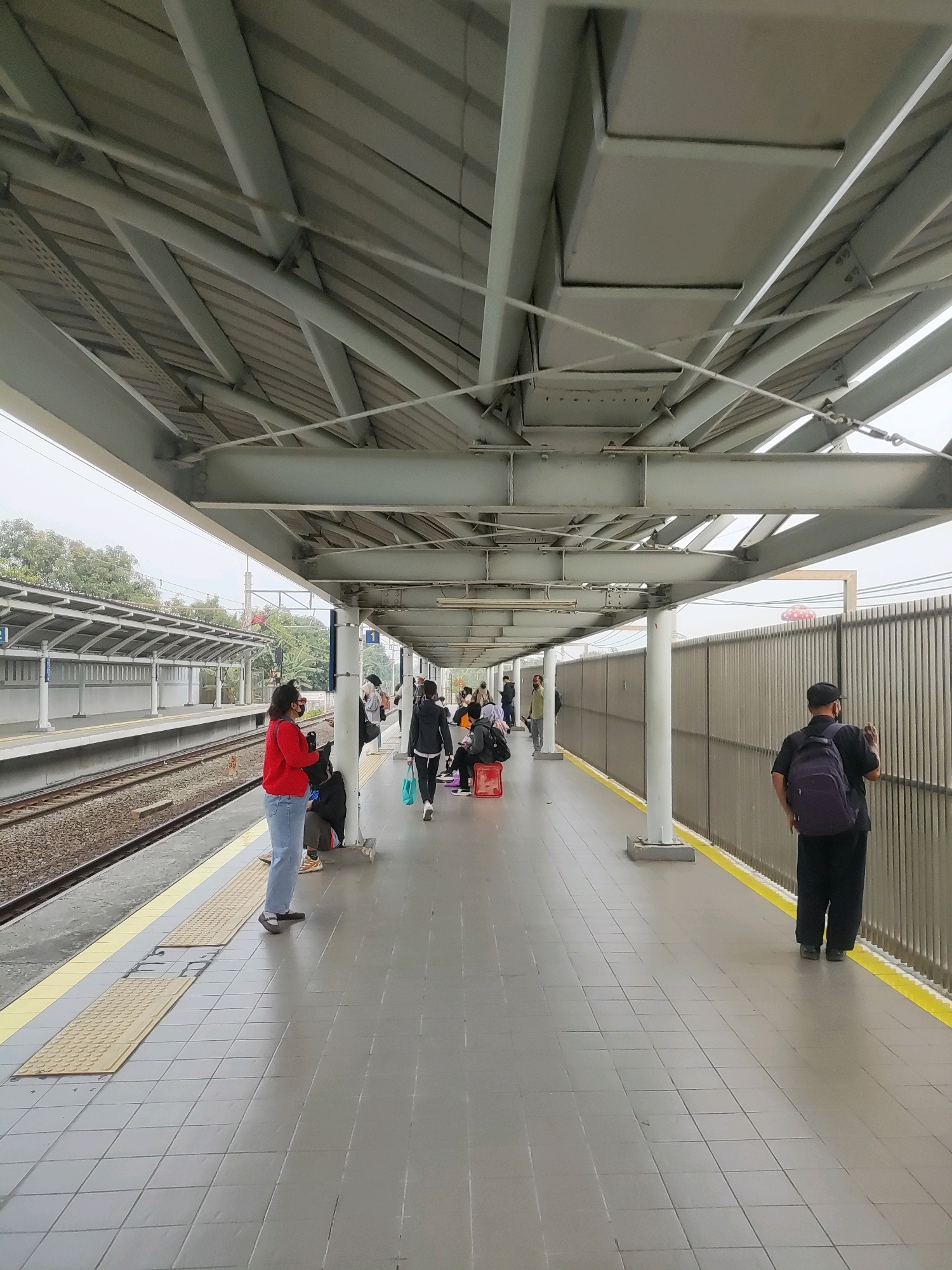 A train station platform in Telagamurni, Indonesia, features a covered walkway with a grey tiled floor and yellow safety lines. Several people are waiting for a train, some seated and others standing. A woman in a red top and jeans stands near the tracks, while a man in a black shirt and pants with a purple backpack faces away from the camera, looking at a fence. The platform is supported by white pillars and a grey metal roof structure. Train tracks run along the left side of the image, and a metal fence lines the right. Trees and a smokestack are visible in the background. The sky is overcast.