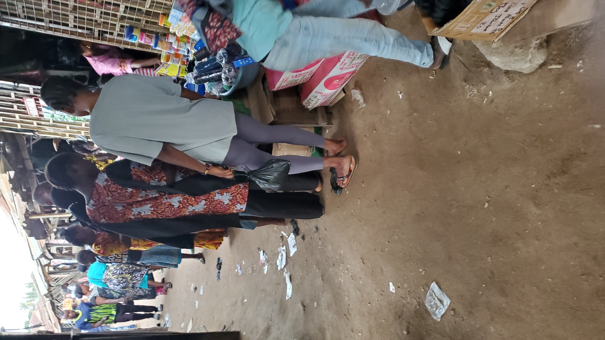 A busy marketplace scene unfolds in Jalingo, Nigeria. Several individuals are visible, primarily women, in various states of activity. Some are standing in what appears to be a queue, while others are engaged in transactions or observing. The ground is a dusty, uneven dirt surface littered with debris, including paper scraps and what appear to be discarded shoe soles.
In the foreground, a person wearing light blue jeans and brown sandals stands with their back to the camera, holding a black plastic bag. To their left, a woman in a patterned orange and black dress is also partially visible. Further back, a person in a light grey t-shirt and purple leggings stands facing away from the camera. A man in a blue and green shirt is visible on the far left, facing towards the marketplace stalls.
The environment suggests a typical outdoor market with makeshift stalls and shelters constructed from corrugated metal and wood. Goods are displayed on shelves and in containers, with visible items including packaged products, plastic containers of various colors, and fabric. The overall impression is one of a vibrant, albeit somewhat disorganized, commercial hub within the city. The lighting indicates it is daytime, with soft, diffused light suggesting an overcast sky or shaded area.