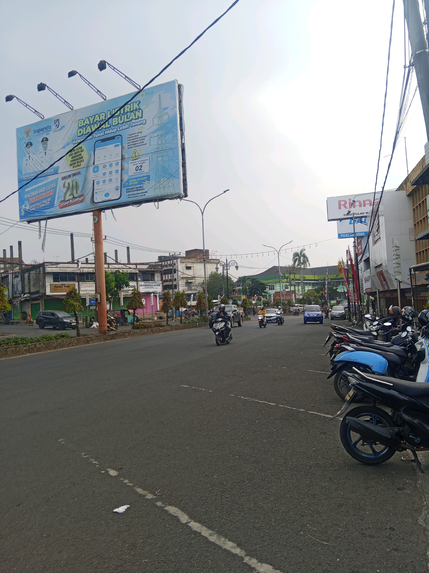 This is an eye-level, wide shot of a street scene in the city of Lubuklinggau, Indonesia. The street is paved and has white lane markings. On the right side of the street, a row of motorcycles is parked, including a blue scooter in the foreground. Across the street, there are several buildings, including storefronts with signs. A large billboard is visible on the left side of the image, advertising electricity bill payments. Several vehicles, including cars and motorcycles, are on the road, and some people are visible riding motorcycles. The sky is overcast. The overall impression is that of a busy urban environment.