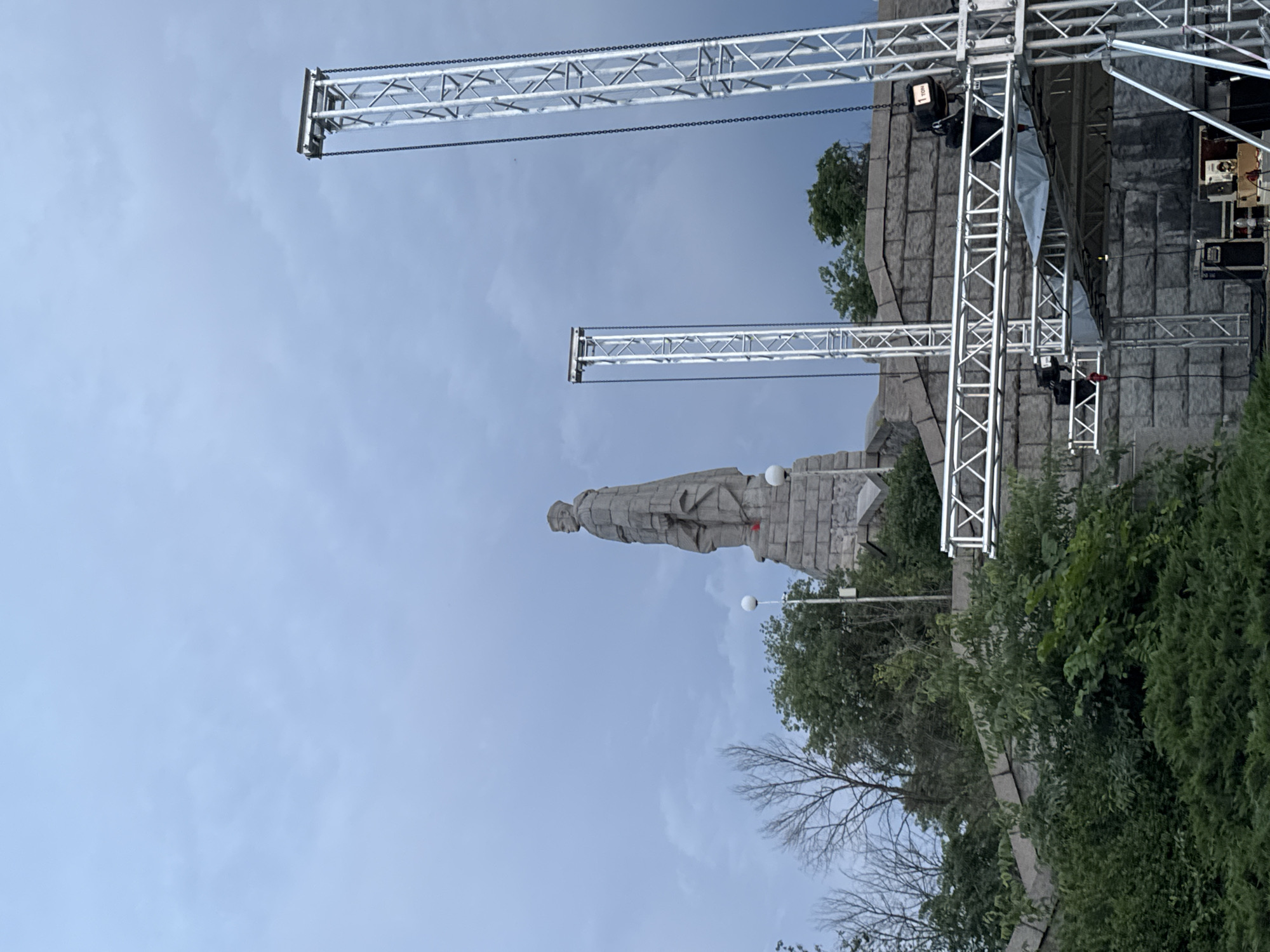 The image captures a significant historical monument juxtaposed with modern event infrastructure. The central subject is a large, imposing grey stone statue of a human figure, appearing to be a soldier, standing on a substantial stone pedestal or base. This monument is likely the Alyosha Monument in Plovdiv, Bulgaria, given the provided location context.
In the foreground and partially obscuring the monument, a complex setup of silver metal truss structures indicates preparations for a large public event, such as a concert or performance. These trusses extend vertically and horizontally, fitted with chains and black lighting fixtures. A partial view of a stage with dark equipment is visible on the right side of the image, further supporting the idea of an upcoming event.
The setting includes lush green trees and bushes surrounding both the monument and the event staging. Two spherical white street lights are also visible near the monument. The sky is a uniform, light blue-grey, suggesting an overcast day, likely late morning or afternoon, providing soft, diffused lighting to the scene.
A notable detail is the text "1 TON" visible on a black box attached to one of the truss structures, indicating its load capacity. The image combines elements of historical significance with contemporary event planning, suggesting a blend of cultural reverence and public celebration or gathering.