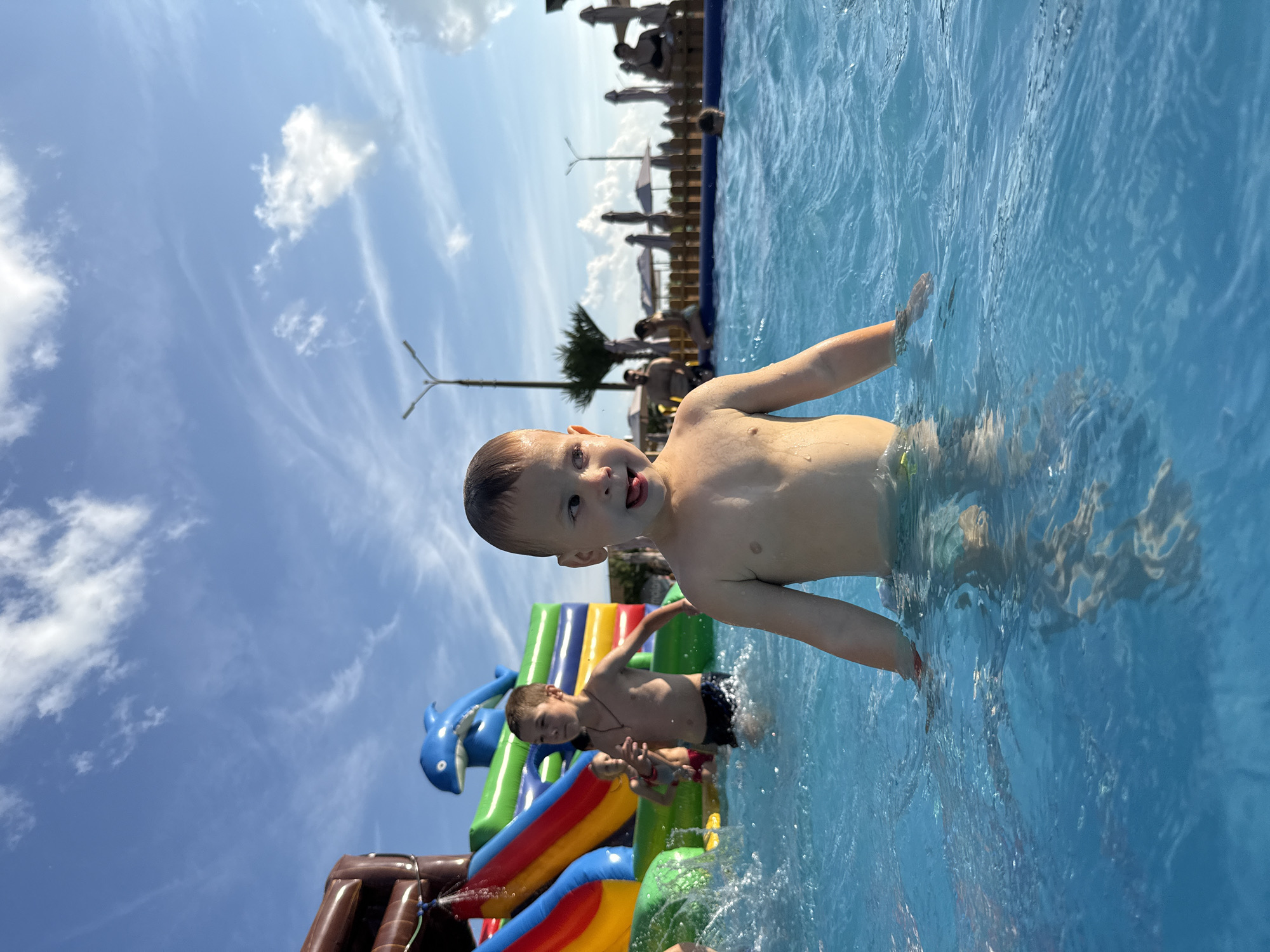 In a bright outdoor scene, a young boy with fair skin and short brown hair is wading in a pool of clear blue water. He is facing to his right and sticking his tongue out. He is wearing a pair of swim trunks that are not fully visible. Behind him, another boy with dark hair is also in the water, wearing swim trunks. Further behind, a colorful inflatable water slide with a blue dolphin on top is partially visible on the left. Other people are visible in the background, some sitting on lounge chairs and others in the water, with umbrellas providing shade. The sky above is a clear blue with scattered white clouds. The location appears to be an outdoor water park or pool area.