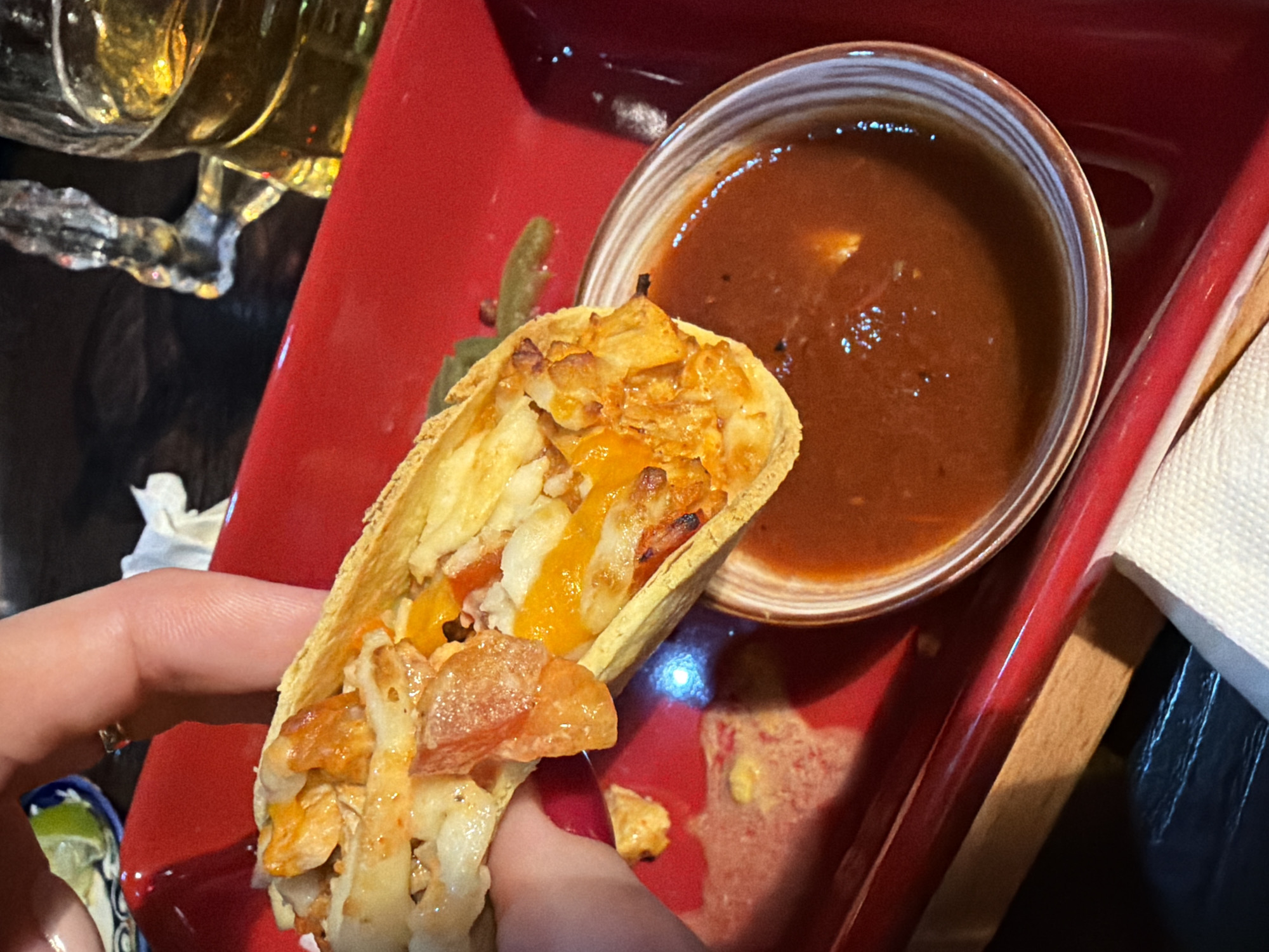The image shows a person holding a taco filled with shredded chicken and tomatoes. Next to the taco, there is a bowl of dark red salsa on a red rectangular plate. In the background, there is a partially visible glass of beer and a lime wedge in a small dish. The setting appears to be a restaurant, likely located in Rostov-on-Don, Russia.