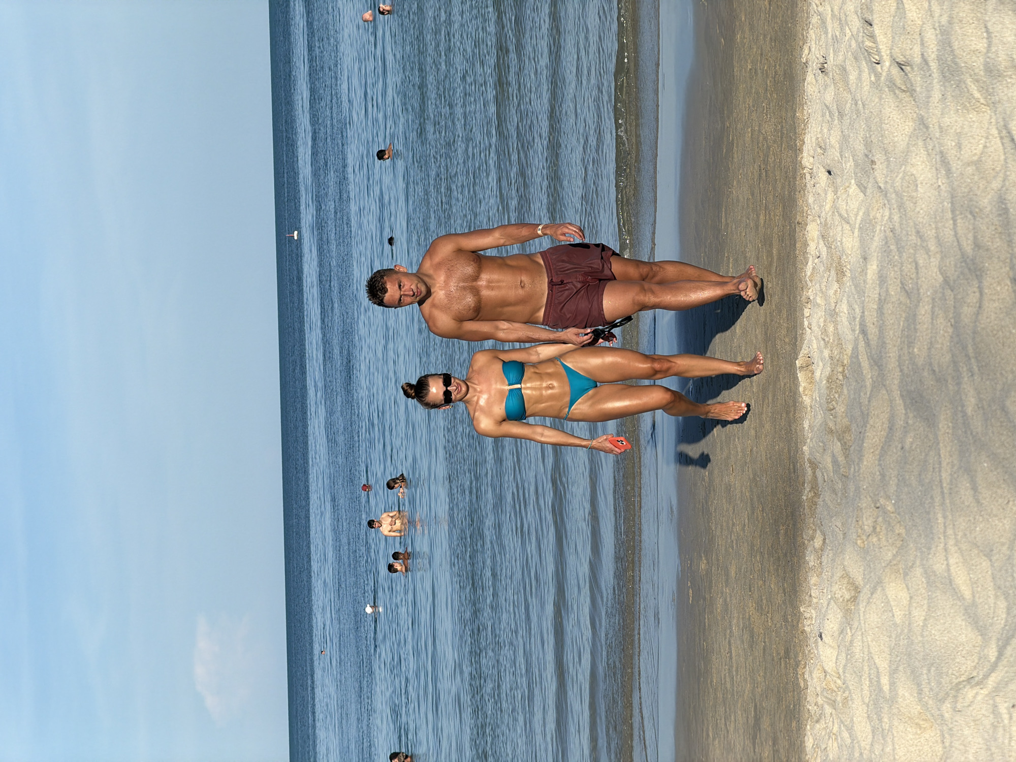 A man and a woman walk side-by-side on a sandy beach toward the viewer. The woman is on the left, wearing a teal bikini, sunglasses, and is holding a red smartphone in her right hand. She has her hair in a bun and is looking forward. The man is on the right, wearing dark red swim trunks, and is holding a pair of sunglasses in his right hand. He is shirtless and looking forward. They are both tanned and appear fit. In the background, the ocean is visible with several people swimming and playing in the water under a clear blue sky with a few wispy clouds. The location is identified as Baan Chaweng, Thailand.