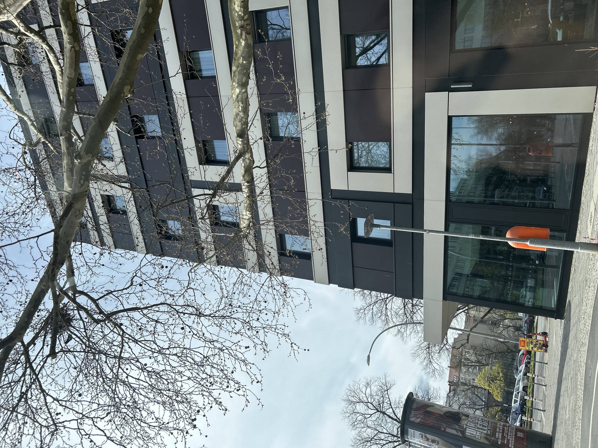 The image shows an upward view of a modern building in Berlin, Germany. The building is tall with a black and white striped facade and numerous windows. A bare tree with numerous thin branches and some seed pods is prominent in the foreground, partially obscuring the building. The sky is overcast.
In the background, across the street from the modern building, are other buildings, trees, parked cars, a scooter, a bus stop, and a light post. A large cylindrical advertising pillar is in the foreground on the right side of the frame.