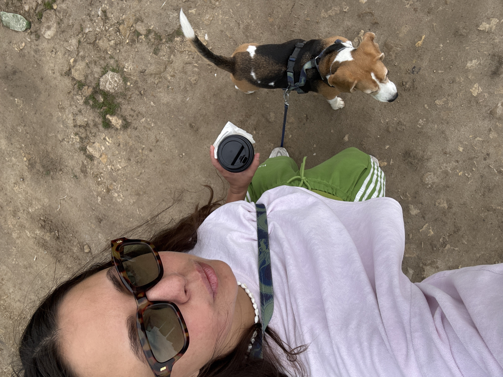A top-down perspective shot depicts a person walking a dog on a dirt path. The person is holding a closed, black to-go coffee cup in their right hand. They wear tortoise-shell sunglasses, a white pearl necklace, a light purple shirt, and green sweatpants with white stripes down the side. The dog, a beagle with black, brown, and white fur, is on a blue leash and wearing a black harness. The ground is dirt with patches of exposed rocks and small bits of vegetation. The location in the city of Kazan, Russia is not determinable from the image.