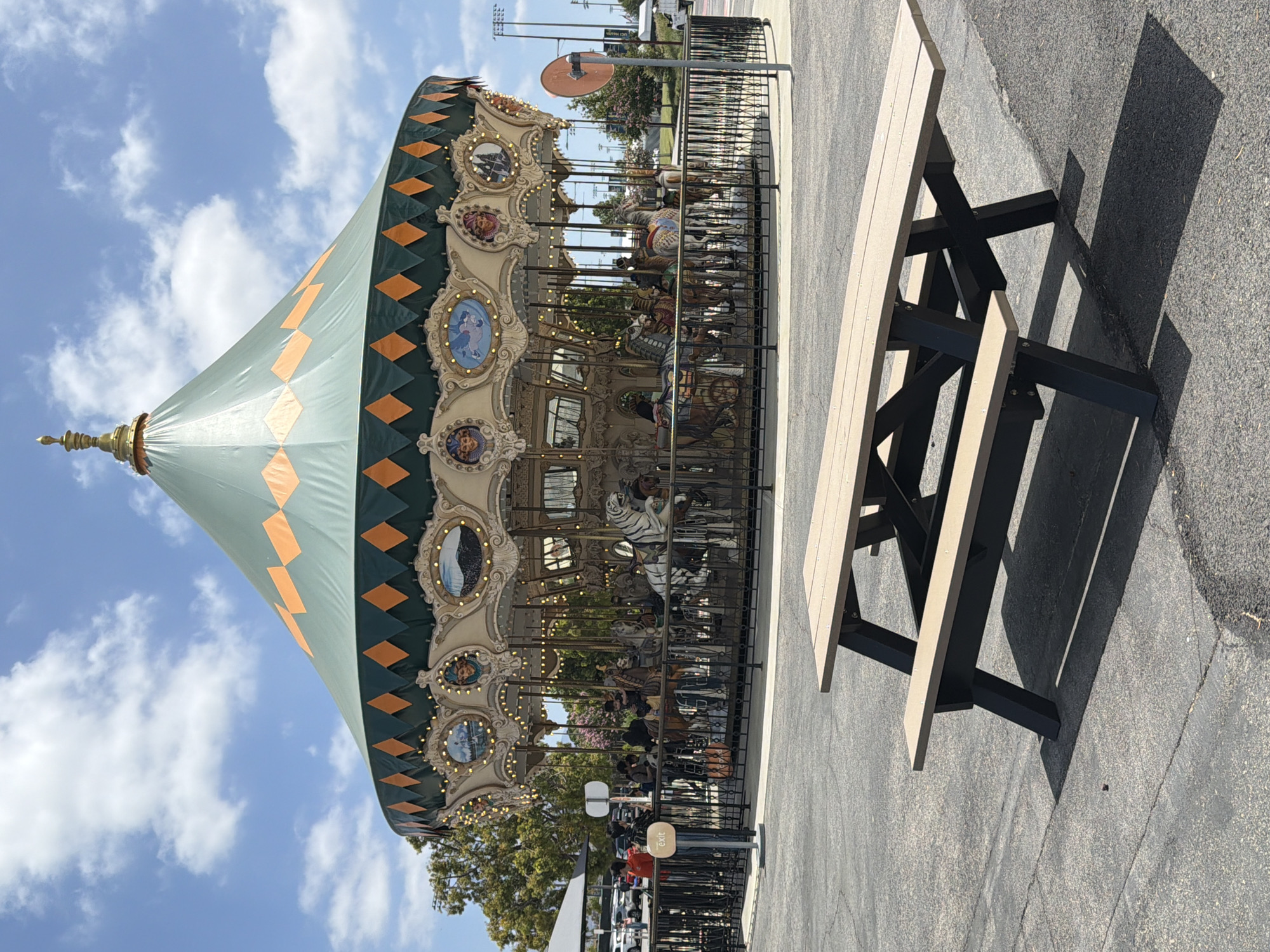 This is a medium shot, eye-level, outdoor, full-color photograph taken on a sunny day with some scattered clouds. In the foreground, a tan and dark brown picnic table casts a distinct shadow on the asphalt ground. Behind the picnic table, a carousel is prominently featured. The carousel has a large, green, conical roof adorned with orange diamond shapes. The carousel is fenced off with black metal railings, and several people can be seen inside and around the carousel, some appearing to be on the rides. The carousel features various horses and other mounts, decorated with ornate details and lights. In the background, trees and a parking lot with cars are visible, suggesting an amusement park or recreational area. The overall impression is a bright, cheerful scene on a typical day. This carousel is located at the Great Park in Irvine, California.