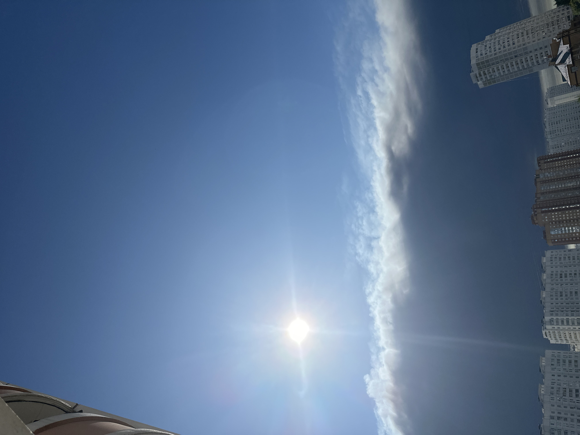The image shows a dramatic sky over the city of Podolsk, Russia. The sky is divided into two distinct layers. The upper portion is a clear, bright blue, illuminated by a strong sun with visible rays and lens flare. Below this, a thick, dark, ominous cloud bank stretches horizontally across the frame. A thin band of lighter, wispy clouds sits at the edge between the bright sky and the dark cloud bank. In the foreground, the tops of several apartment buildings are visible against the stormy sky, suggesting an urban environment. The buildings are of various heights and architectural styles, with some appearing modern and multi-story. The overall impression is one of an approaching storm or a dramatic weather event occurring over the city.