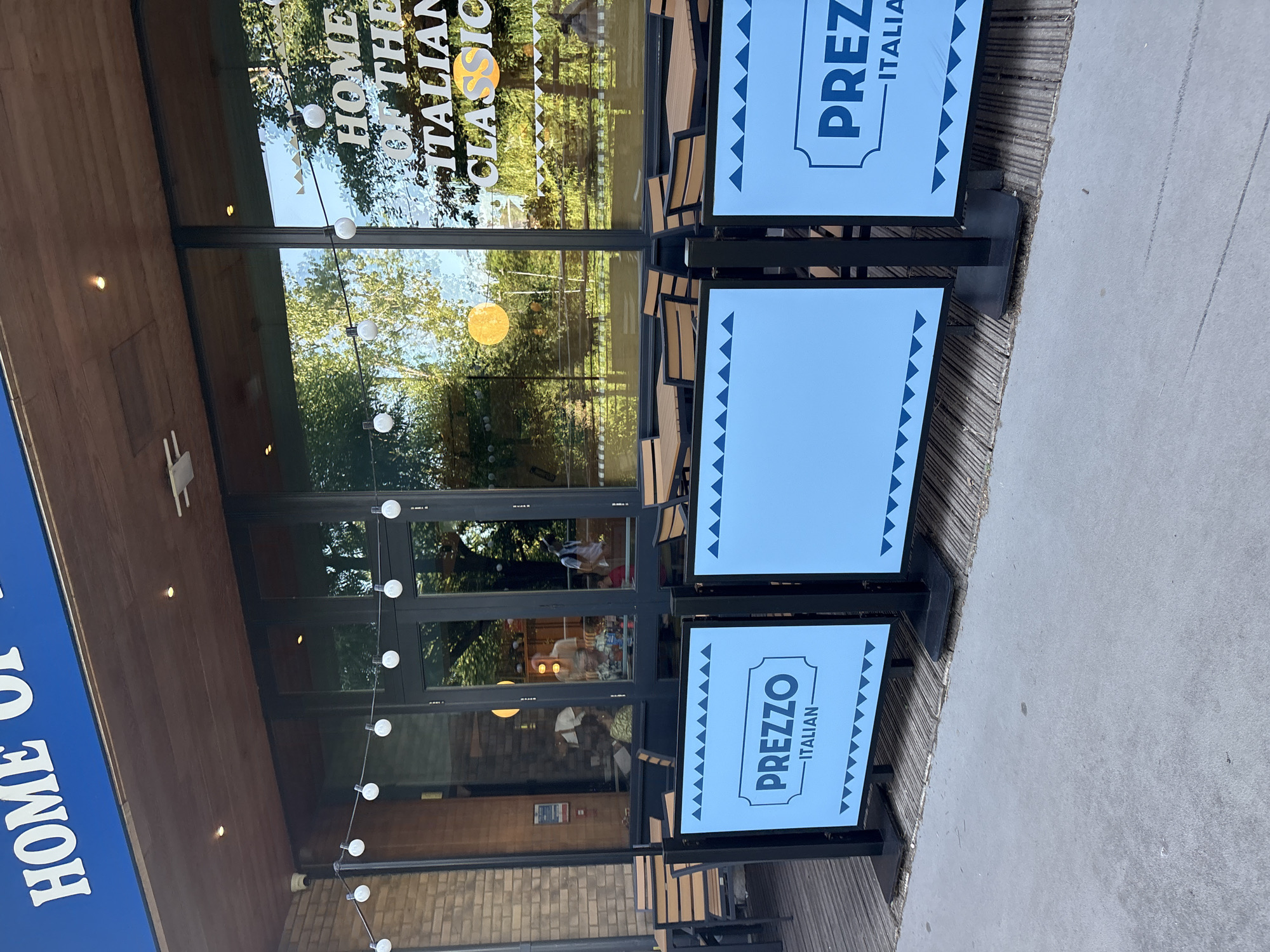 The image depicts the exterior of a restaurant called "PREZZO ITALIAN." The scene is set outdoors, with a modern, clean aesthetic. Two digital signs are prominently displayed, advertising "PREZZO ITALIAN" with a light blue background and bold, dark blue lettering within a bordered frame. The signs have a decorative border of small blue triangles.
The restaurant entrance features large glass windows and doors, allowing a view into the interior where tables and chairs are visible. The interior appears to be well-lit, with some reflections of trees and the sky on the glass. There are string lights with round bulbs hung outside the entrance, suggesting it might be evening or an overcast day. Wooden paneling is visible on the building facade, adding a touch of warmth to the contemporary design.
On the left side of the image, a blue sign with white lettering reads "HOME OF THE ITALIAN CLASSIC." This suggests the restaurant has a long-standing reputation or specializes in traditional Italian cuisine. The overall impression is that of a welcoming and stylish dining establishment. The image's orientation is vertical, and the lighting indicates it was likely taken during daylight hours.