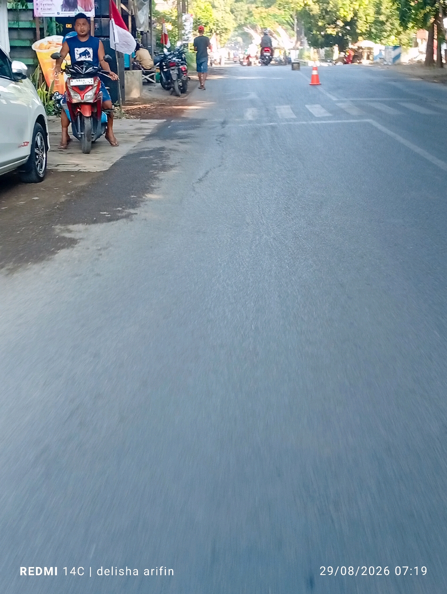 This image captures a street scene in Plinggisan, Indonesia, on August 29, 2026, at 07:19. A young man wearing a blue sleeveless shirt with a bull graphic and blue shorts is seated on a red scooter. He appears to be looking forward, with his hands on the handlebars. The scooter is parked on a paved surface next to a sidewalk. Behind him, there are several motorcycles parked, and a white and red Indonesian flag is visible, suggesting it might be a public area or near a shop. Further down the street, a pedestrian in a dark shirt and shorts is walking away from the camera. The road ahead has a pedestrian crossing marked with white lines and a traffic cone. Trees and buildings line the right side of the street, indicating a developed urban environment. The lighting suggests it is early morning. The bottom of the image contains text identifying the device as "REDMI 14C" and a name "delisha arifin".