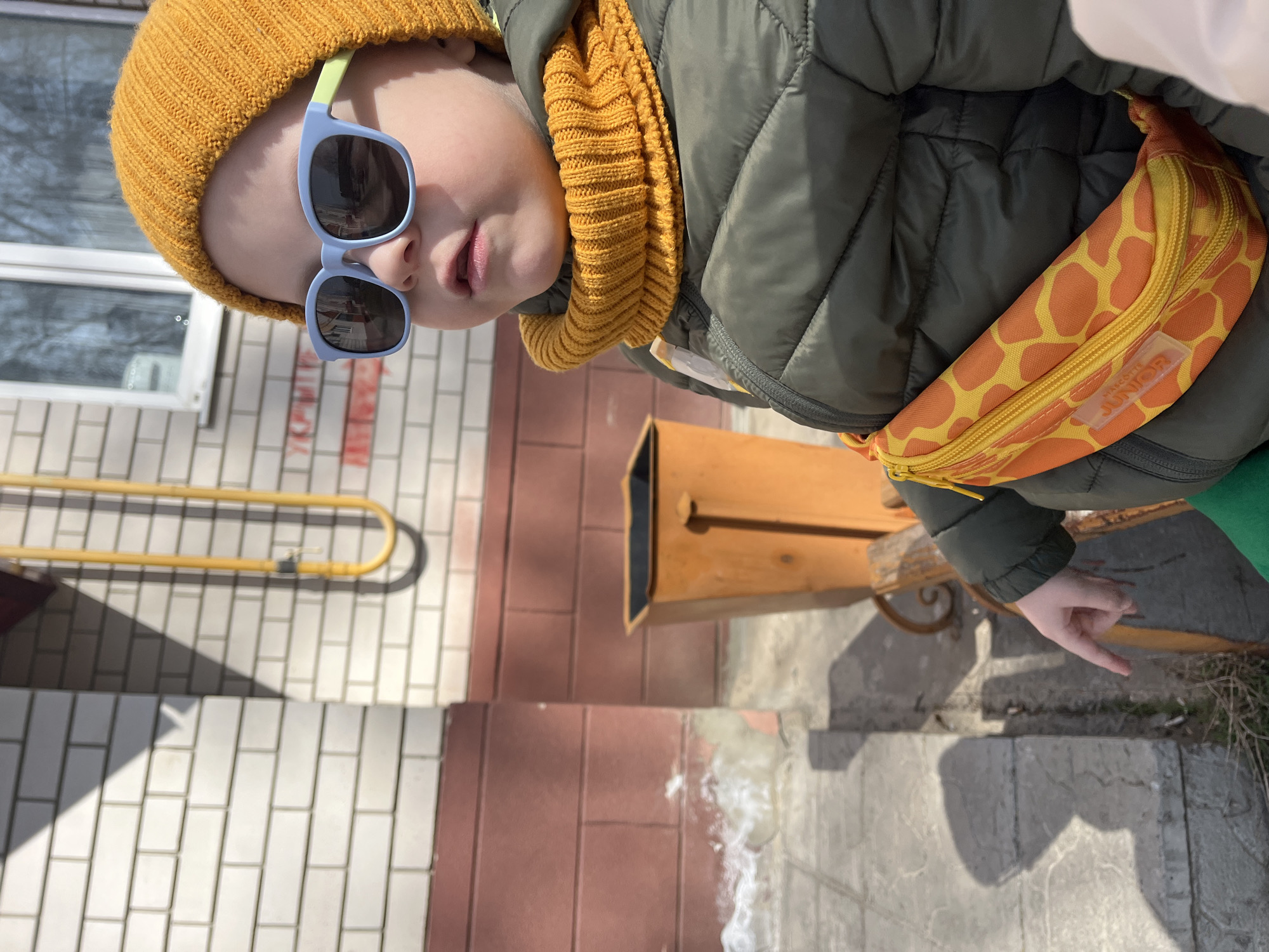 A child wearing a yellow beanie, sunglasses, a yellow scarf, and a green jacket with a giraffe-print fanny pack stands outside. The child points toward the ground. A bench and brick wall are visible in the background. The scene is outdoors, likely in Usman, Russia.