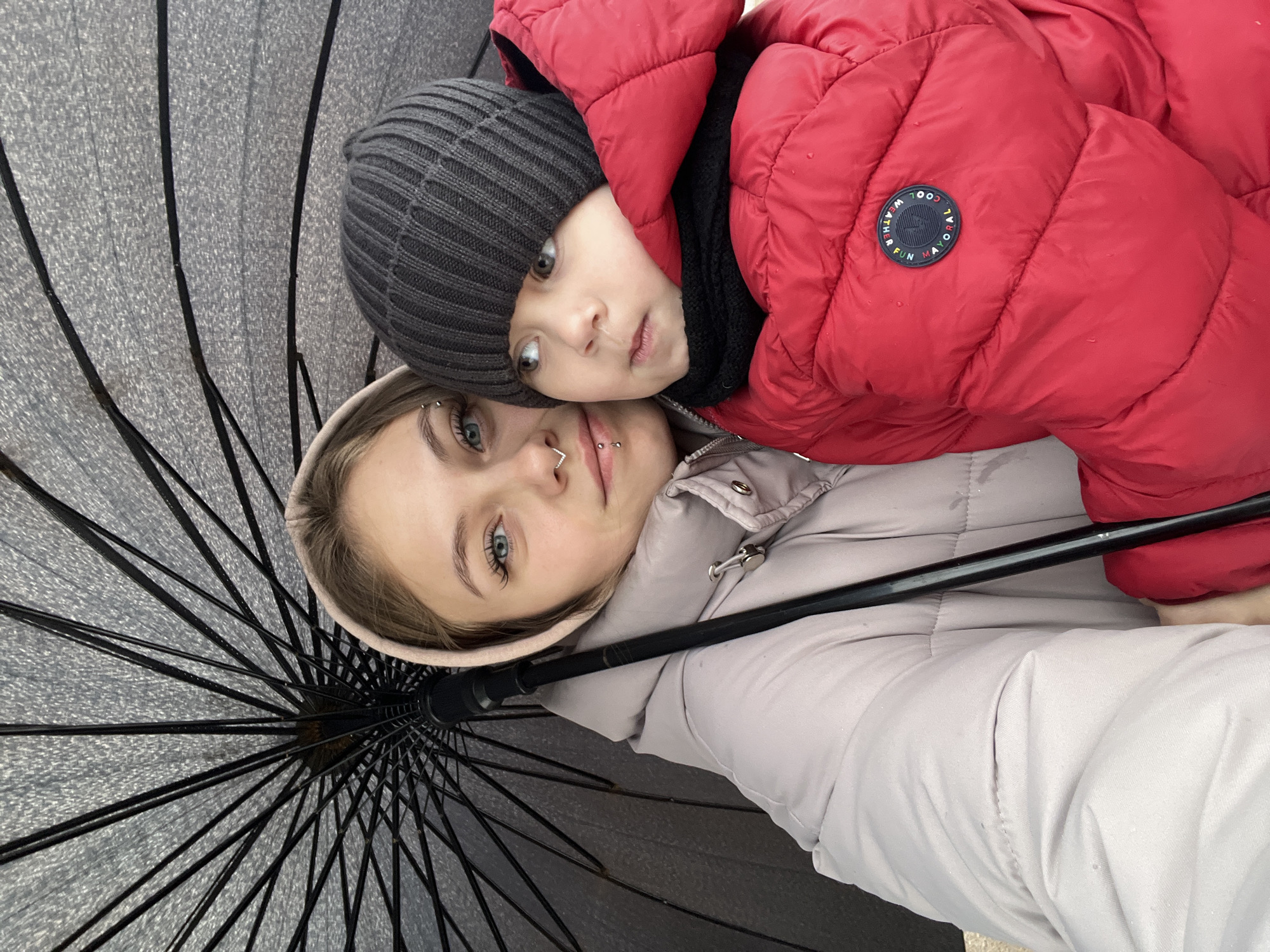 A selfie features a young woman and a child, likely a parent and child, under an umbrella. The woman is on the bottom, wearing a light pink hooded jacket, with facial piercings on her eyebrow and nose. The child is above her, wearing a red puffer jacket and a grey knit hat. The umbrella is open, filling a portion of the frame. They appear to be outdoors, potentially in Фоки, Russia, though the specific location within the city is not discernible. They are interacting by being close together in the selfie.