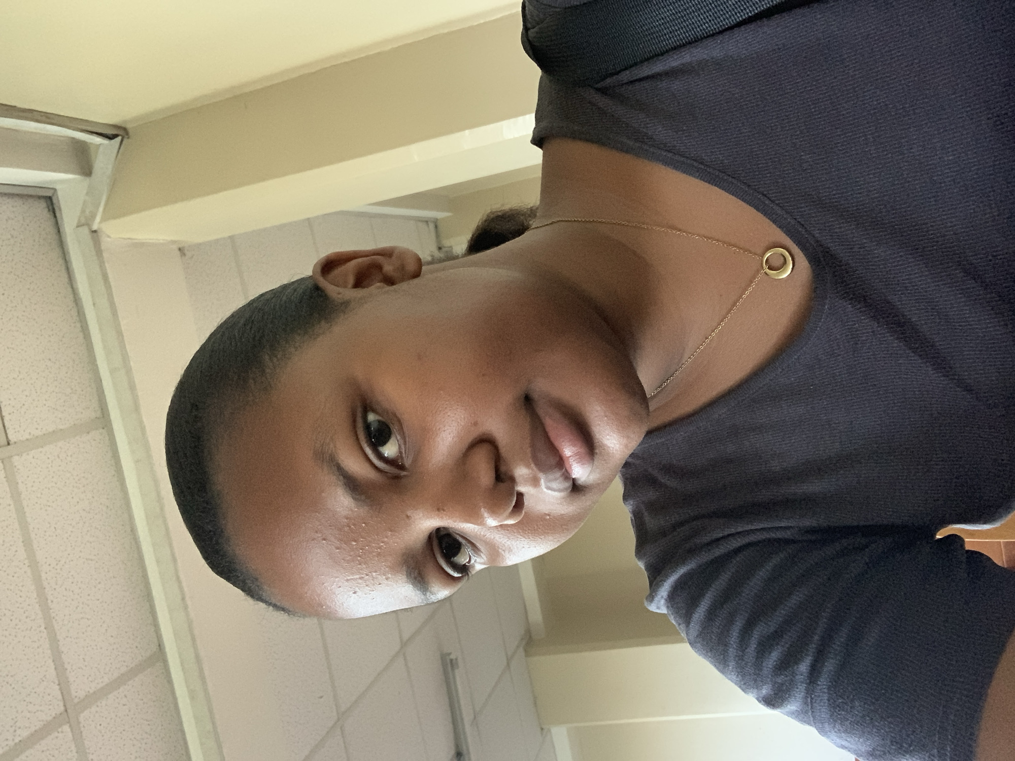 A close-up, rotated portrait-style selfie captures a young person with dark skin looking directly at the camera. Their dark hair is pulled back, revealing a clean hairline. The subject wears a dark-colored, possibly ribbed or textured, short-sleeved top, and a delicate gold chain necklace with a small, circular gold pendant rests on their chest. A black backpack strap is partially visible over their shoulder.
The background indicates an indoor setting, likely a commercial or institutional building. Light-colored walls and a suspended ceiling with square tiles are visible, along with a portion of a window or door frame. The lighting suggests artificial indoor illumination or diffused natural light. The overall mood is neutral. No text is visible in the image. The location context provided is Dar es-Salaam, Tanzania.