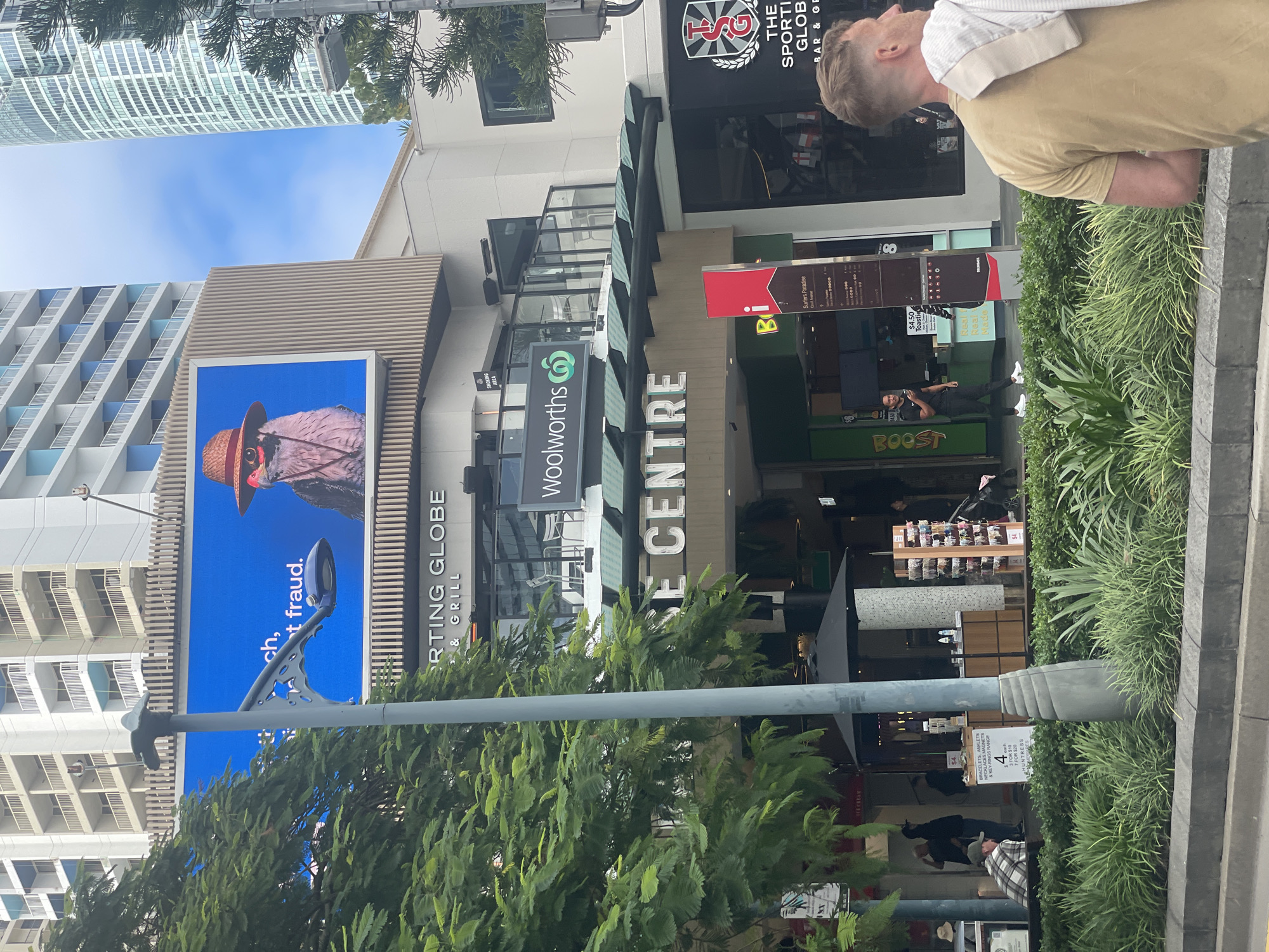 An outdoor, eye-level, medium shot captures a street scene in Surfers Paradise, Australia. In the background, a large digital billboard displays a close-up of a bird wearing a straw hat and sunglasses, with text that partially reads "ch, t fraud." Below the billboard, the facade of a building features prominent signage for "SPORTING GLOBE BAR & GRILL" and "WOOLWORTHS." The lower portion of the image shows lush green foliage and a street-level view of people walking and sitting. A grey lamppost with a decorative base stands in the foreground, slightly off-center. The sky is partly cloudy.