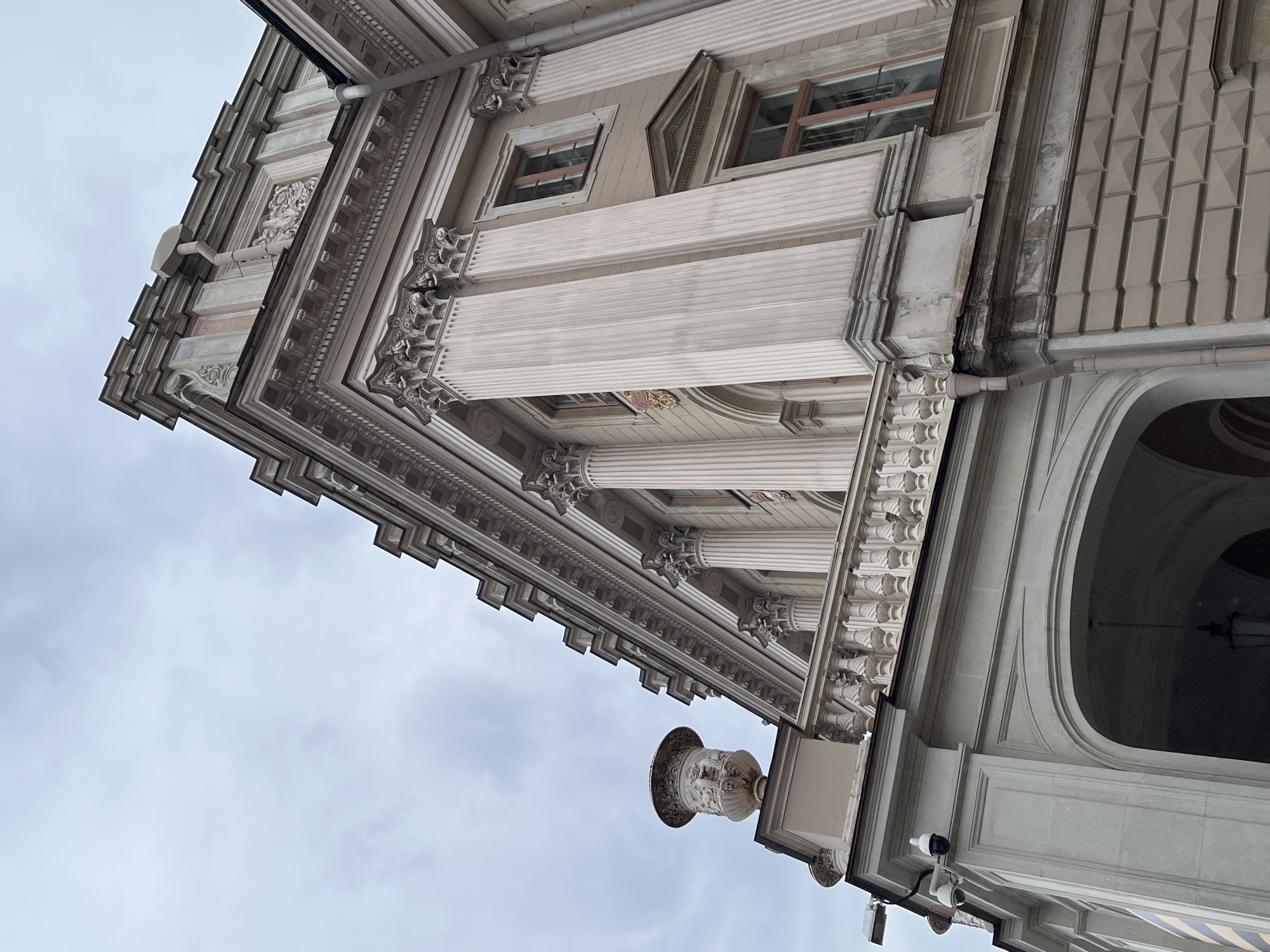 Here's a concise description of the image:
This is a low-angle shot of a section of the Winter Palace in Saint Petersburg, Russia. The architecture displays ornate detailing, including columns, window frames, and decorative elements. Visible architectural features also include a cornice, arched entryway, and an exterior wall. The angle of the shot and overcast sky contribute to the building's aesthetic. Surveillance cameras are attached to the exterior structure.
