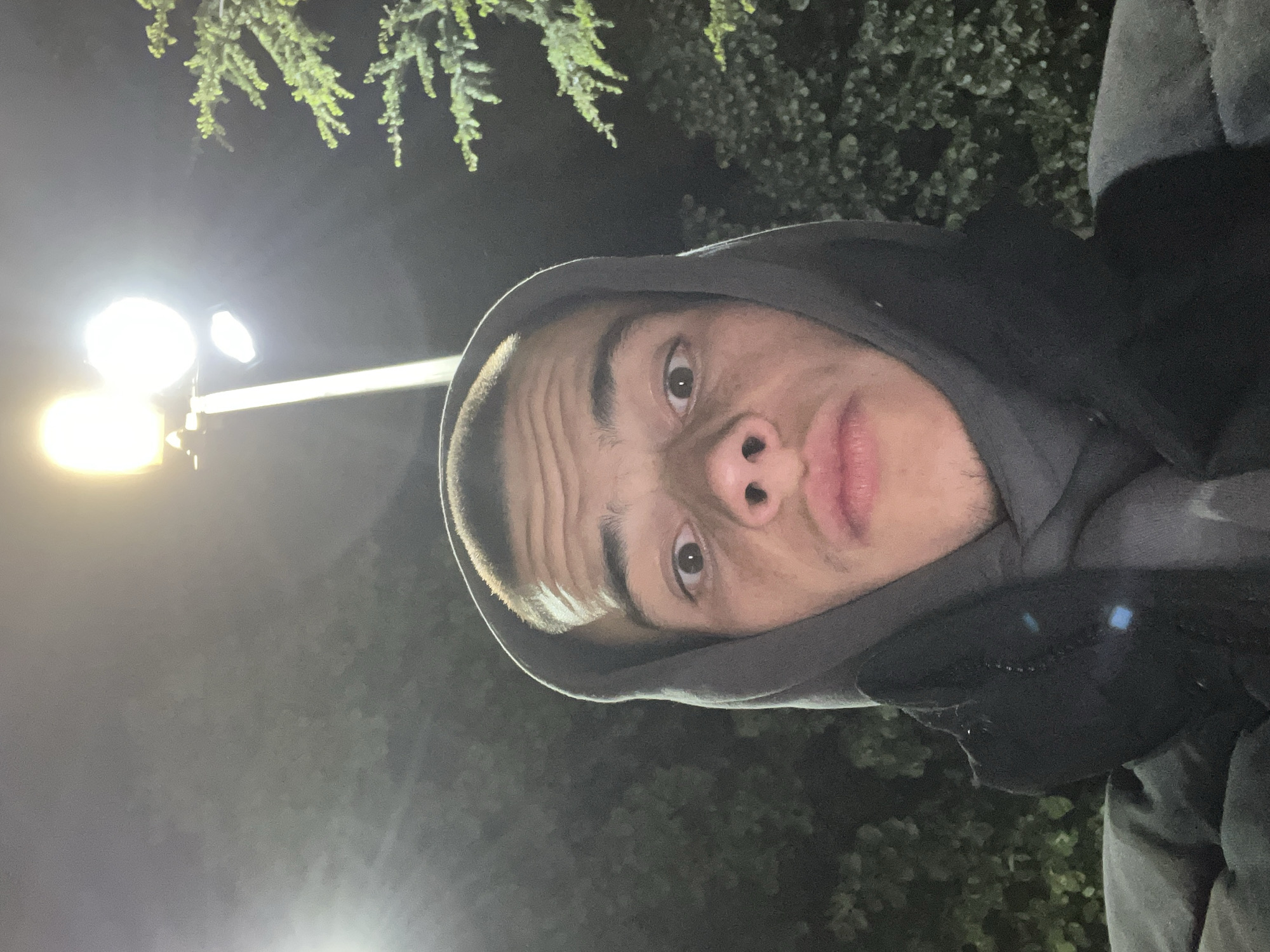 A selfie of a young man with light skin and short hair, wearing a hooded sweatshirt, is taken outdoors at night. A street light is visible in the background. Vegetation is also visible in the background. The location is not evident from the image.