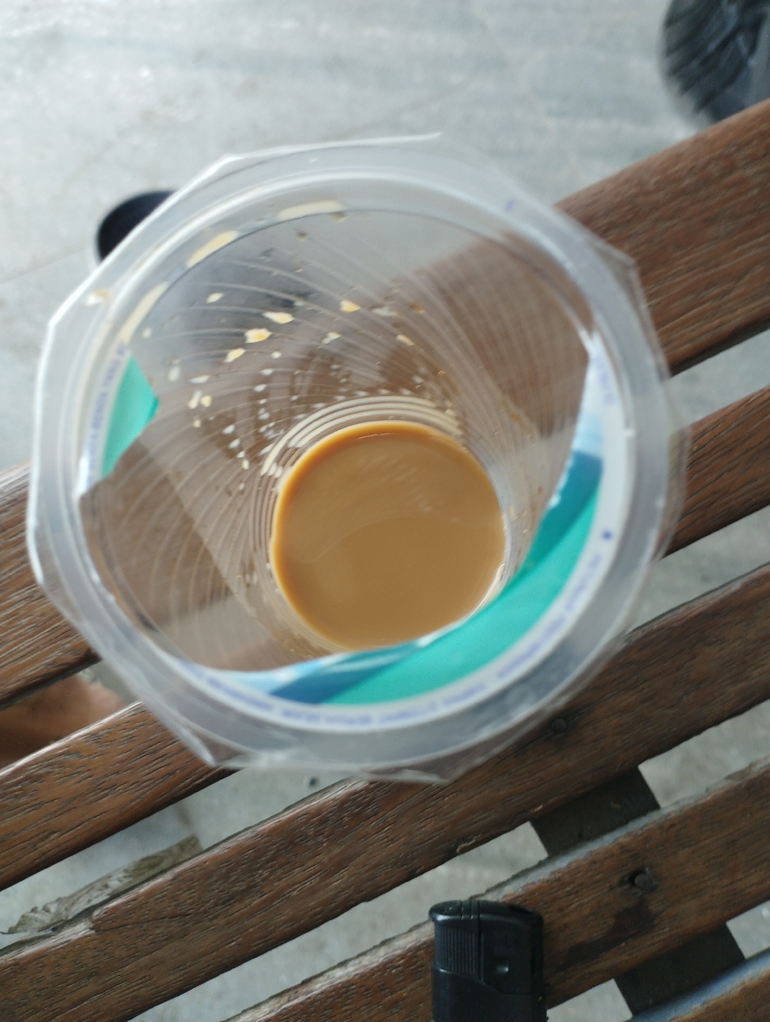 The image shows a partially full, clear plastic cup containing a light brown liquid, likely coffee or tea with milk, sitting on a wooden bench. The bench is composed of multiple parallel wooden slats. There are remnants of what may be dried milk or coffee residue on the inside walls of the cup. Partially visible in the lower right corner is a black lighter. The location is potentially Lhokseumawe, Indonesia.