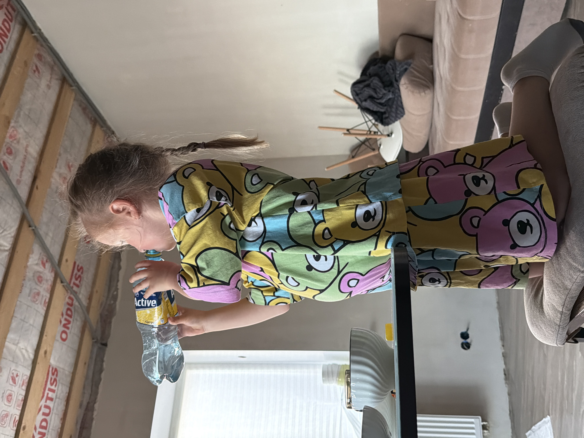 An indoor, vertically oriented image, taken in Chaikovsky, Russia, features a young child drinking from a plastic water bottle. The child, likely a girl with light brown hair styled in two braids, is wearing a vibrant, long-sleeved shirt and pants adorned with a pattern of cartoon bears in shades of yellow, pink, blue, and green. She holds a partially full bottle of water with a yellow label that clearly displays the brand name "Active" and part of a number "86".
The scene presents a mix of finished and partially constructed elements within a room. To the left, a wall under renovation is visible, covered with white insulation material branded "ONDUTISS" and reinforced with wooden studs. In the background to the right, a light-colored sofa or armchair and a small white chair with wooden legs are present, with a dark piece of clothing draped over the latter. Part of an adult's legs, clad in light-colored socks, can be seen resting on the sofa on the far right. A white radiator is partially visible in the lower right. Natural light streams in from a window with blinds or shades in the mid-ground, indicating it is daytime. A white fan or air purifier on a dark stand stands in the foreground. The child appears focused on her drink.