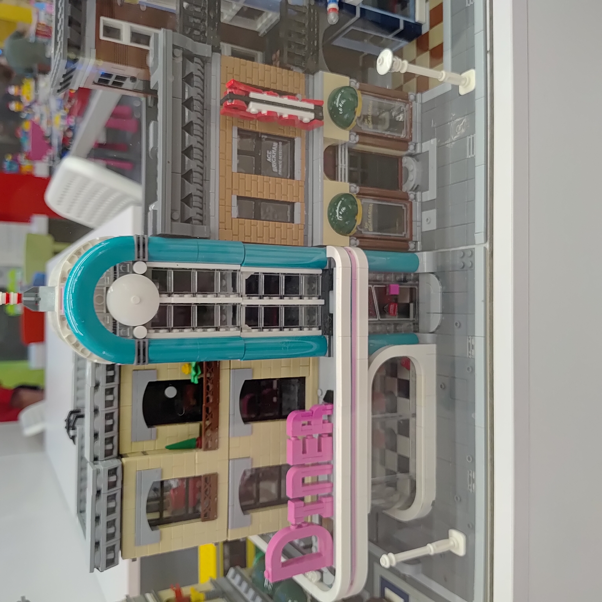 The image shows a close-up, eye-level view of a Lego model of a diner and adjacent buildings. The diner is prominently featured in the foreground, with large pink "DINER" lettering on its facade. The building has a retro, 1950s aesthetic, with a teal archway and a large circular element above the entrance. To the right of the diner is another building with a brick facade, a red vertical sign that appears to say "ACE BRICKMAN PRIVATE DETECTIVE," and windows with checkered patterns. There are also two green, dome-shaped light fixtures on this building. To the left of the diner, part of another building is visible, with yellow brickwork and windows. The model is set on a gray Lego street base with checkered sidewalk tiles and a white lamppost. In the blurred background, other Lego structures and some out-of-focus people and furniture are visible. The image does not provide any information about the location in the city of Ufa, Russia.
