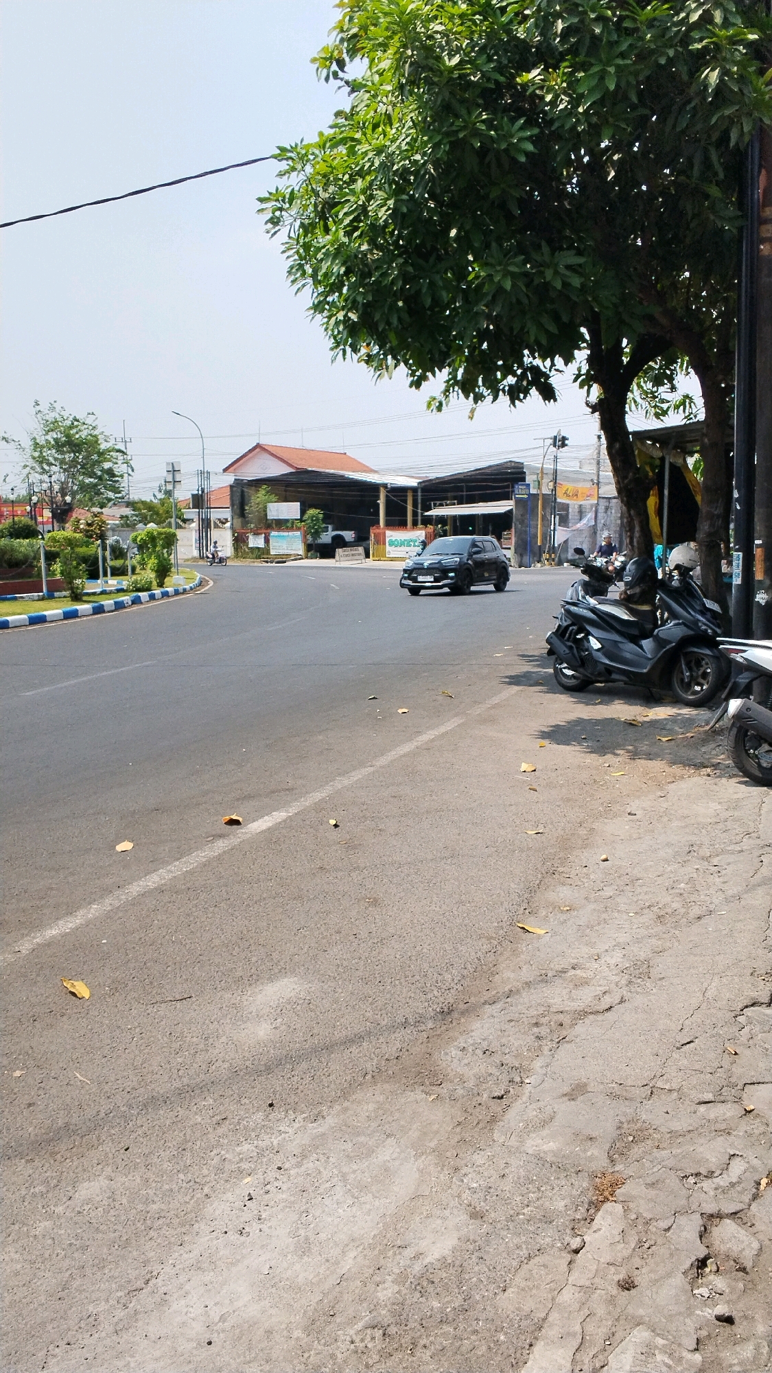 The image captures a street scene in Banjaragung, Indonesia. A black Honda Brio is navigating a curve on an asphalt road. Several motorcycles are parked on the side of the road, with one black scooter prominently featured in the foreground. Green trees and some buildings are visible in the background. The sky is clear and bright, suggesting a sunny day. The road appears to be well-maintained, with white lane markings. The area seems to be a mix of residential and commercial establishments.