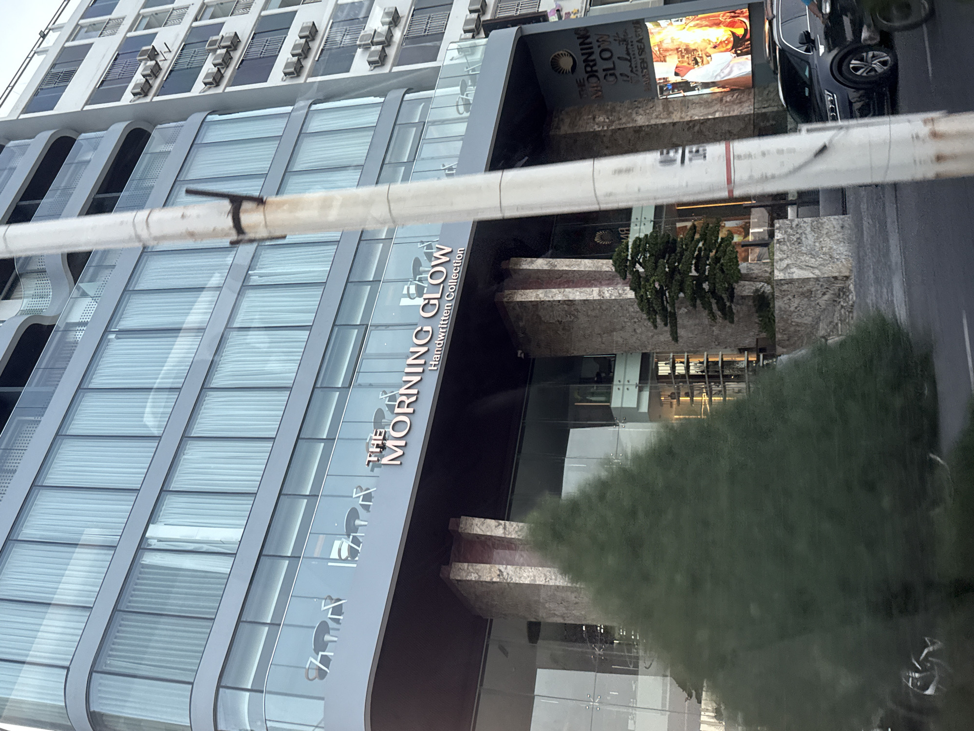 The image shows a tall modern building with a facade of glass windows and white curtains. The building has a sign that reads "THE MORNING GLOW Handwritten Collection." In front of the building, there is a large green conical tree, and to the right, a black SUV is parked on the street. A tall white utility pole is in the foreground, obscuring parts of the building. The scene appears to be a street view in a city, likely in Bac Nha Trang Ward, Vietnam, given the context provided. The lighting suggests it might be daytime, but the sky is overcast.