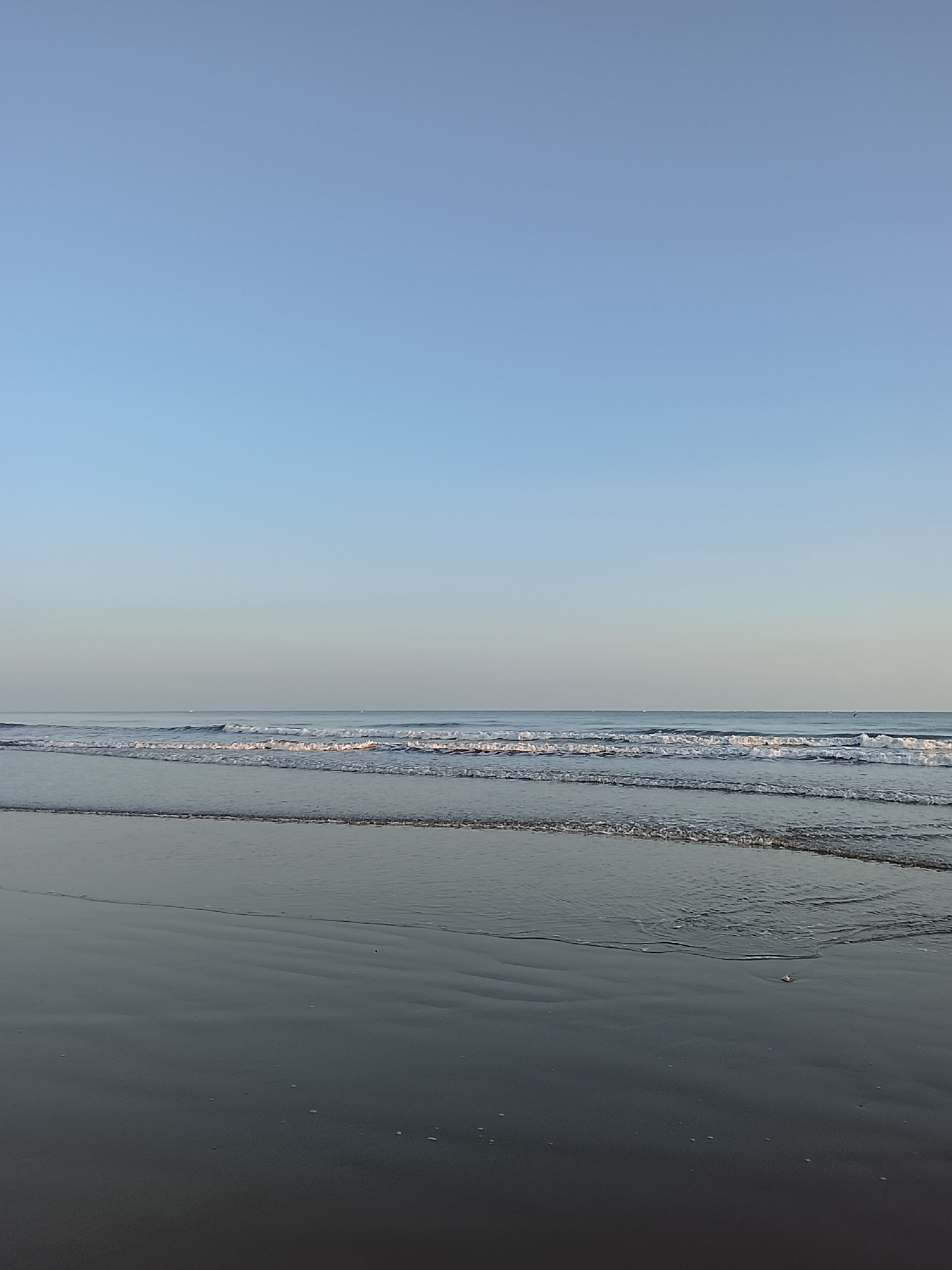 A serene beach scene unfolds under a clear, pale blue sky. The horizon is a faint line where the sky meets the ocean, with a subtle gradient in the sky suggesting the early morning or late afternoon light. Gentle waves are rolling onto a sandy shore. The sand itself is dark and appears wet, reflecting the muted light of the sky. Several lines of small, foamy waves break and recede, creating a textured pattern on the water's surface. In the distance, a few faint, dark specks, possibly distant boats or birds, are visible on the water. The overall impression is one of tranquility and natural beauty.
