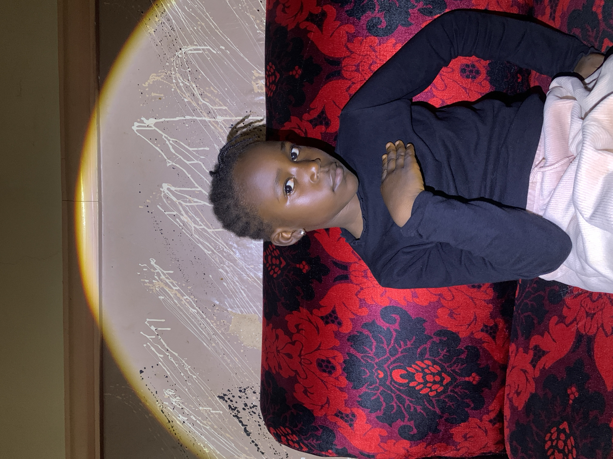 A young Black child with braided hair lies on a patterned couch, their body mostly covered by a black long-sleeved shirt and light-colored pants. The couch has a vibrant red and black floral design. The child's head is tilted, and their gaze is directed towards the viewer. One hand rests on their chest, while the other is tucked beneath their head.
To the left of the couch, a wall is visible. It appears to be a light, neutral color and is decorated with white and black paint splatters, resembling abstract art. A distinct beam of warm, yellow light falls across the wall from the left, creating a gradient of illumination and casting a shadow. This suggests the light source is positioned to the left of the frame, possibly a lamp or a flashlight.
The overall scene is indoors, likely in a home or living space in Barakin Sabawa, Nigeria. The lighting and the splattered paint on the wall might indicate a creative or perhaps a slightly unkempt environment. The child's expression is neutral, perhaps curious or thoughtful. There is no visible text in the image.