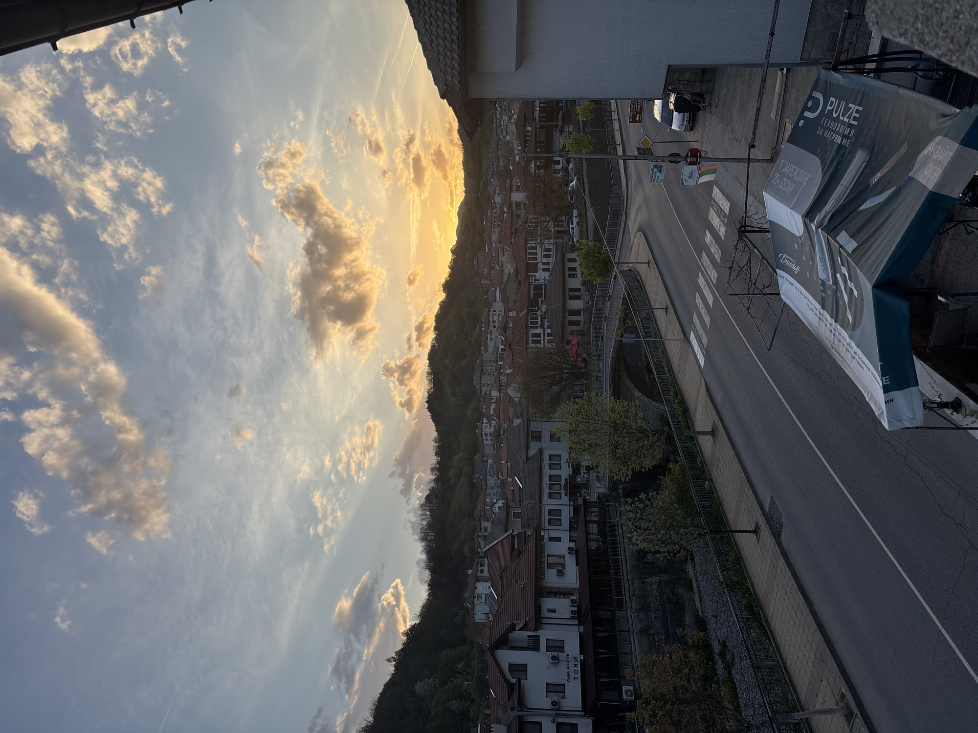 This image presents a serene evening vista over a town, likely Zlatograd, Bulgaria, set against a backdrop of a forested mountain. The perspective is from an elevated position, possibly a balcony, looking down onto the town and a street.
The time of day is sunset, with the sun casting a warm, golden glow across the horizon, illuminating scattered stratocumulus clouds in shades of orange and yellow. The sky transitions to a soft blue higher up, with some wispy clouds.
The scene features a dense, green mountain covered in trees, which forms the natural boundary of the town. The town itself consists of numerous buildings with white or light-colored facades and prominent red-tiled roofs, characteristic of traditional architecture in the region. Some buildings display balconies and air conditioning units.
In the mid-ground and foreground, an asphalt road with clear white lane markings and a prominent pedestrian crosswalk curves through the urban landscape. Streetlights and various road signs line the thoroughfare. A silver car is parked on the side of the road. On the right side of the foreground, a large, dark-colored banner or awning is visible, likely belonging to a commercial establishment.
Visible text on the large banner includes "PULZE" (repeated), "ТЕХНОЛОГИЯ ЗА НАГРЯВАНЕ" (Technology for heating), "1 ЗАРЕЖДАНЕ 25+ СЕСИИ" (1 charge 25+ sessions), "ОСКВАЙТЕ СКОРО", and "Davidoff," suggesting an advertisement for a tobacco product. A sign on one of the buildings clearly states "КЪЩА ЗА ГОСТИ ТОНИ" (Guest House Toni). Another distant street sign shows "РЕЦЕ."
The overall atmosphere is tranquil and calm, with no specific human activity evident, highlighting the peaceful transition from day to night over this Bulgarian town.