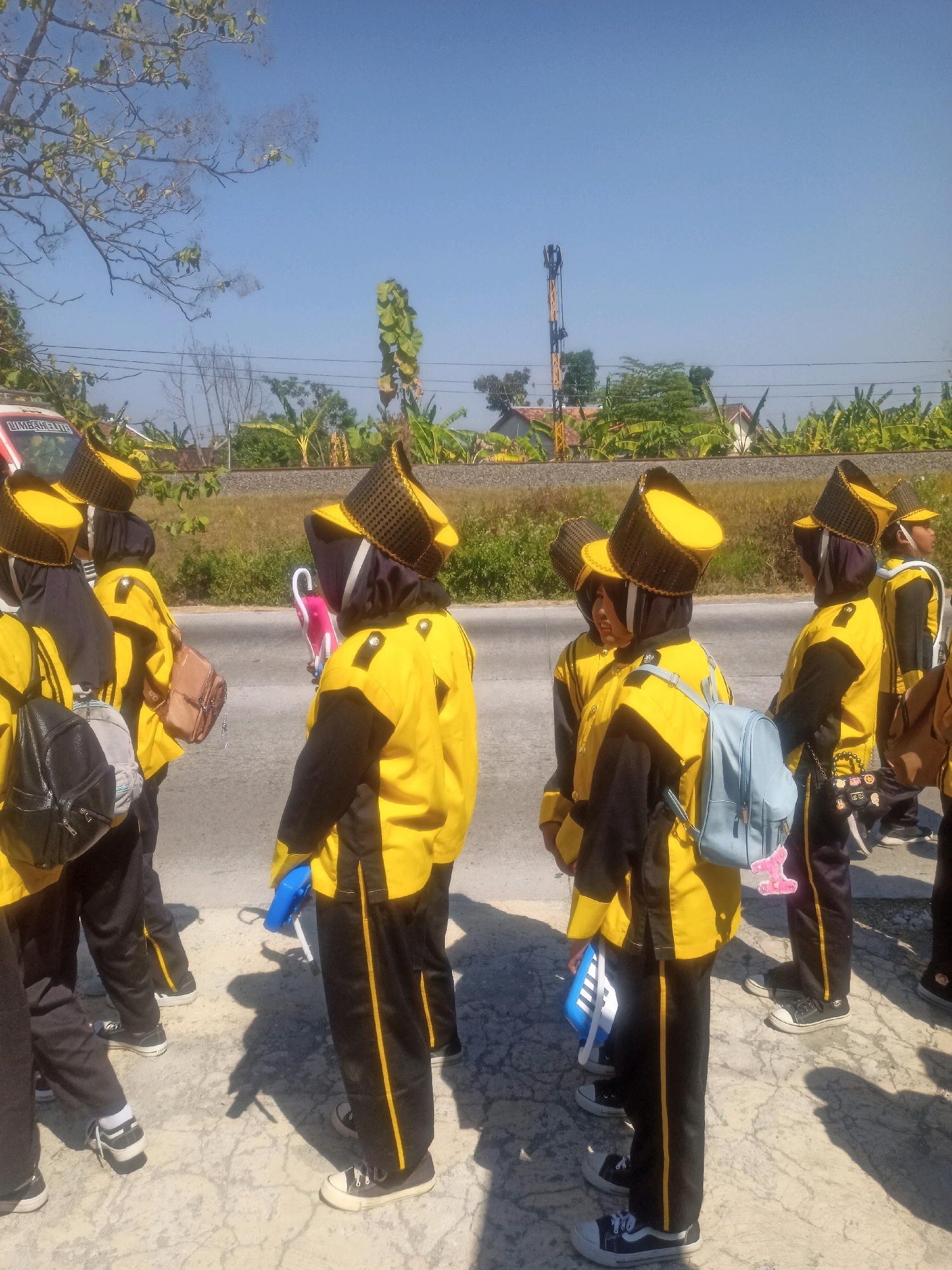 In Sumberlawang, Indonesia, a group of individuals, likely a marching band or color guard, stand on a paved surface beside a road. They are all wearing matching yellow jackets with black sleeves and trim, black pants with yellow stripes, and distinctive tall, cylindrical black and yellow hats. Several individuals have backpacks, and one person holds a pink object, possibly a flag or prop. The background features trees, a cell tower, and what appears to be a railway line. The sky is clear and blue, indicating a sunny day.