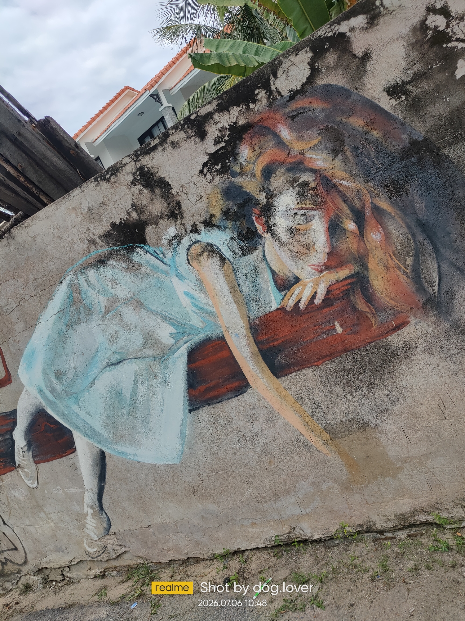 A mural depicts a young woman with her eyes closed, resting her head on a reddish-brown surface. She wears a light blue dress and white shoes. Her left arm extends downward, and her right arm is bent, supporting her head. The mural is painted on a weathered, concrete wall, showing signs of age and grime. In the background, a modern building with an orange tiled roof and banana leaves are visible, suggesting a tropical environment. The photograph was taken in Phan Thiết, Vietnam, as indicated by the metadata.